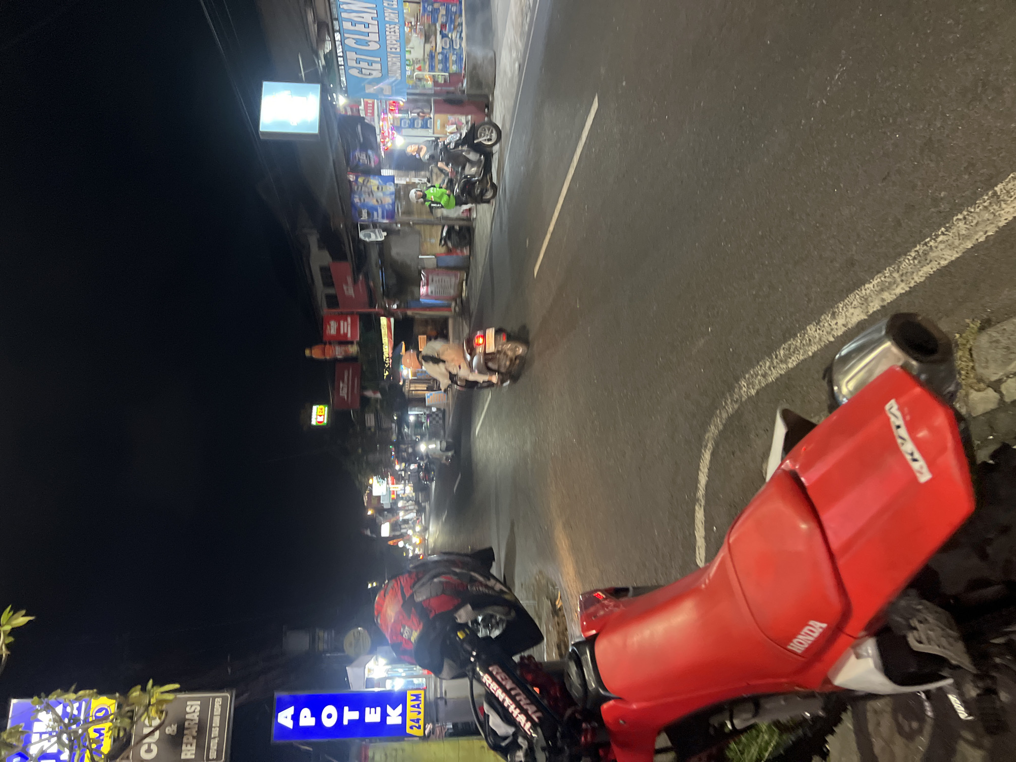 This is a nighttime, eye-level, medium shot of a street scene in Denpasar, Indonesia. The foreground is dominated by the rear section of a red and white Honda dirt bike, its seat displaying the "HONDA" logo. To the left, the handlebars of the dirt bike are visible, adorned with a red and black helmet and a "RENTHAL" branded grip. The street itself is paved and appears wet, with white lane markings.
The background depicts a commercial street with various businesses. On the left, a blue vertical sign reads "APOTEK" (pharmacy) with "24 JAM" (24 hours) underneath. Adjacent to it, a black sign with white lettering states "CL & REPARASI" (cleaning and repair services). Further down the street, shops with illuminated signs line both sides. Several people are present. Two individuals are on scooters: one in a green jacket is stopped near a laundry business, and another person is riding a scooter away from the camera. The overall environment suggests a bustling urban area at night.