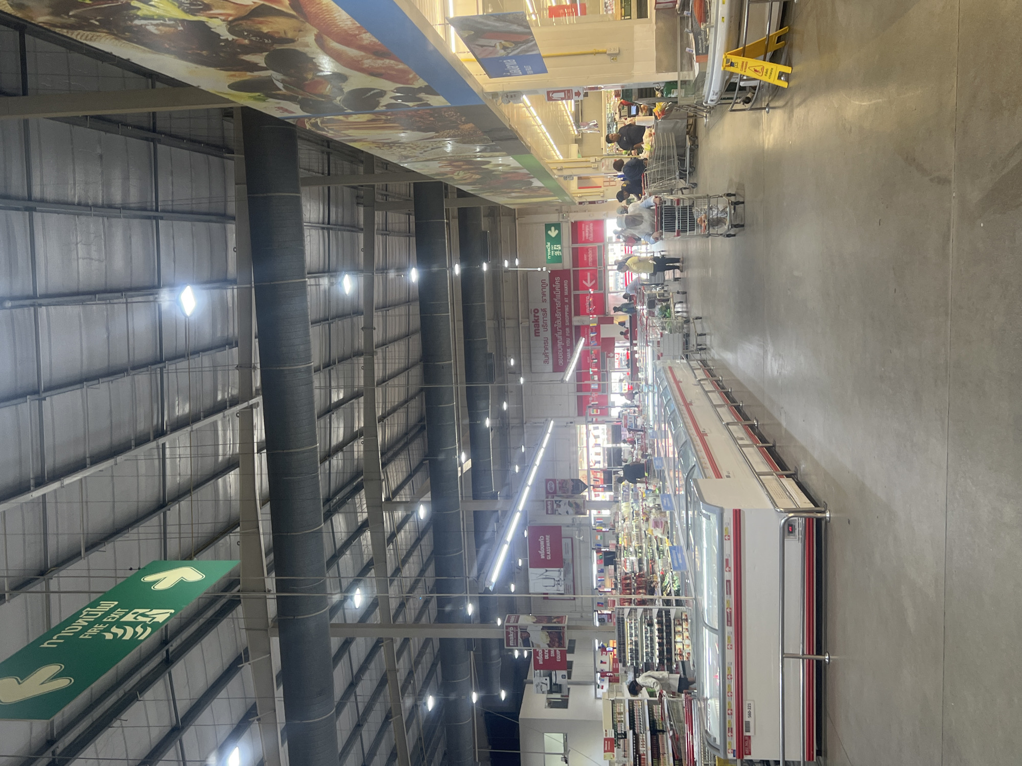 This is a photograph taken inside a Makro wholesale store in Ko Samui District, Thailand. The perspective is from a ground-level view looking down a wide aisle. The store has a high ceiling with exposed metal beams and ductwork, illuminated by fluorescent lights and spotlights. On the left side of the image, there are rows of refrigerated display cases, some with glass lids. Further back, shelves stocked with various products are visible.
In the center and right of the image, shoppers are seen pushing shopping carts. People are dressed casually in t-shirts and shorts. Some are browsing shelves, while others are moving through the aisle. The floor is concrete. There are signs hanging from the ceiling, including directional arrows and product category signs in Thai and English, such as "FIRE EXIT" and "GLASSWARE". A large mural or advertisement featuring food is visible on the right side, running along the top of a shelving unit. A yellow wet floor sign is positioned on the right side of the aisle. The overall impression is a busy retail environment.
