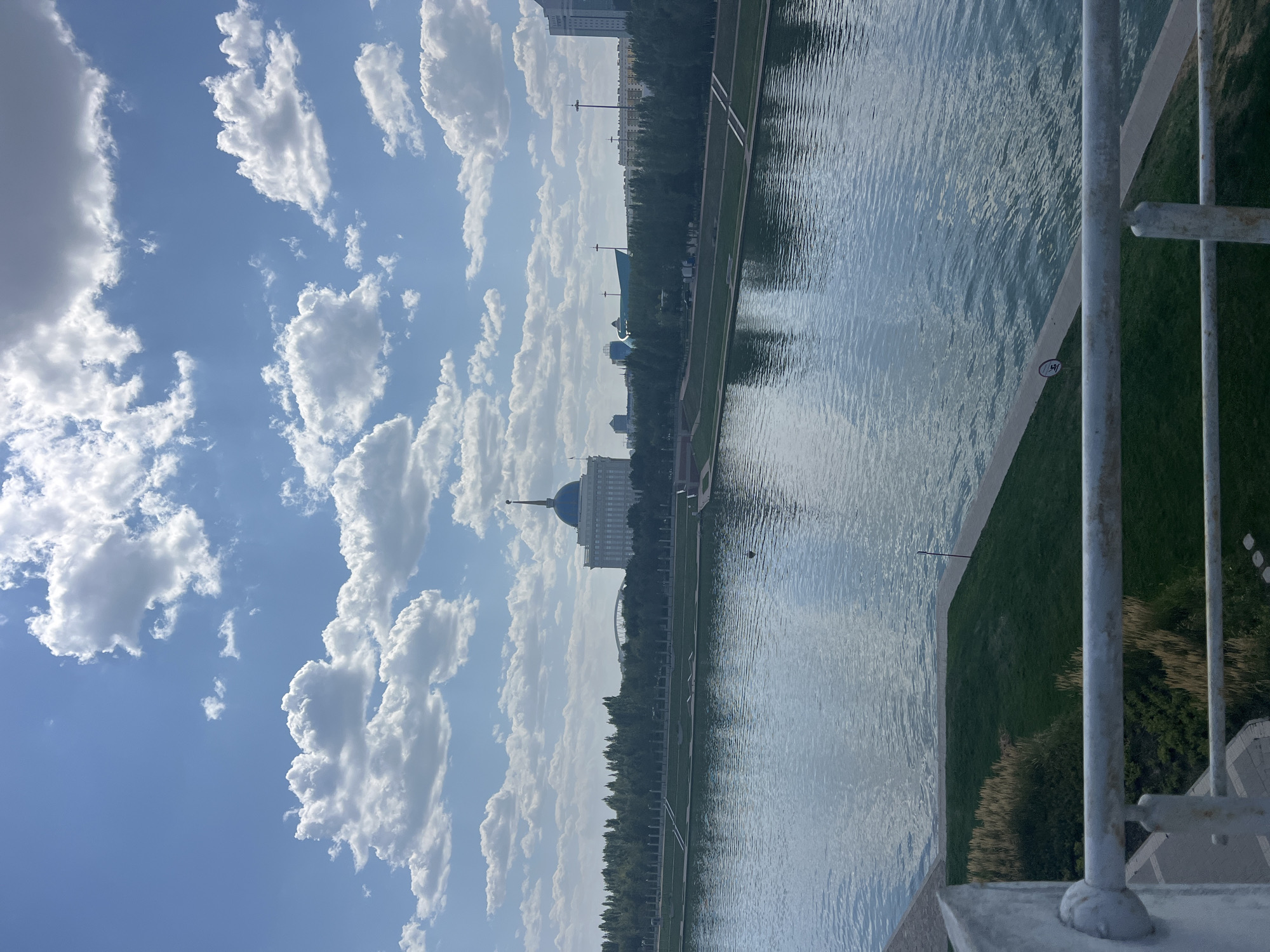 A wide-angle, eye-level outdoor shot captures a scenic view of a city waterfront on a bright, cloudy day. In the foreground, a white metal railing of a bridge or elevated walkway partially frames the scene. Beyond the railing, a section of manicured green grass and a paved pathway slopes down towards a body of water. The water, calm and reflecting the sky and surrounding structures, occupies the middle ground and reflects the overcast sky.
Across the water, a tree-lined embankment forms the backdrop. Beyond the trees, several prominent buildings are visible. The most striking structure is a large, white building with a distinctive blue domed roof topped with a spire, identified as the Ak Orda Presidential Palace. To the right of the palace, other modern urban buildings can be seen, including a distinctive blue, modern architectural structure. The sky above is a vibrant blue, scattered with numerous fluffy white cumulus clouds. A single small, dark object, possibly a buoy or a small boat, is visible on the water. The overall impression is one of urban development integrated with natural elements and architectural landmarks.