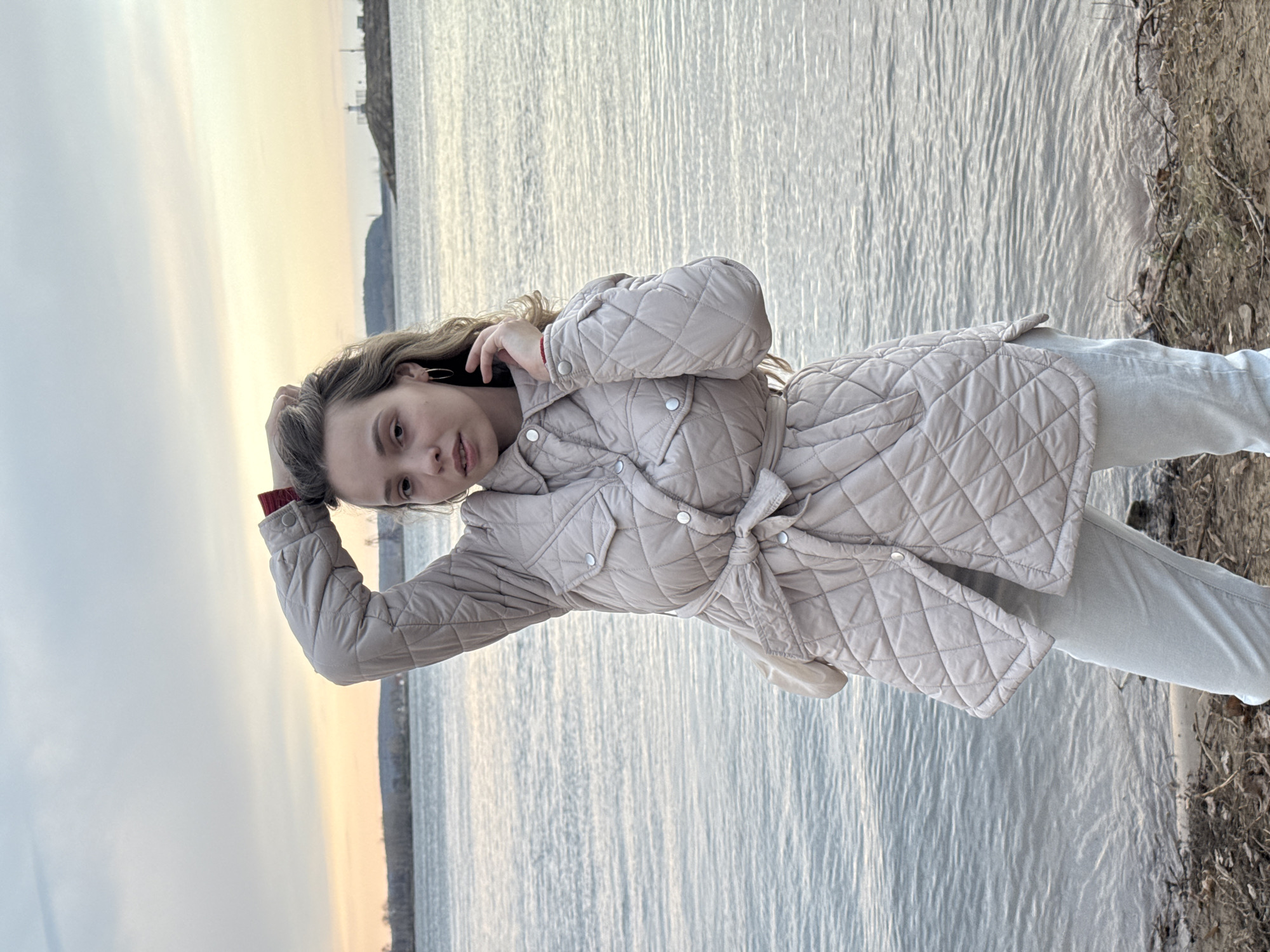 A young woman stands outdoors, posing beside a large body of water, likely the Kama River in Chaikovsky, Russia. She has long, wavy, light brown hair and looks directly at the camera with a calm, neutral expression. She wears a light beige or cream-colored quilted jacket that is tied at the waist, featuring silver buttons and a subtle red cuff visible on her left arm. Her left hand is raised to her head, casually touching her hair. She is also wearing light-colored pants.
The setting places her on a narrow, earthy bank or shoreline on the right side of the frame. The vast expanse of water, with gentle ripples reflecting the soft light, stretches across the mid-ground. In the distance, a faint shoreline with what appears to be trees or structures is visible on the far side of the water. The sky, seen in the upper left, displays the warm, muted tones of either sunrise or sunset, transitioning from a light grey-blue to a pale orange-yellow hue near the horizon, indicating the "golden hour." The overall atmosphere is tranquil and serene. There is no visible text in the image.