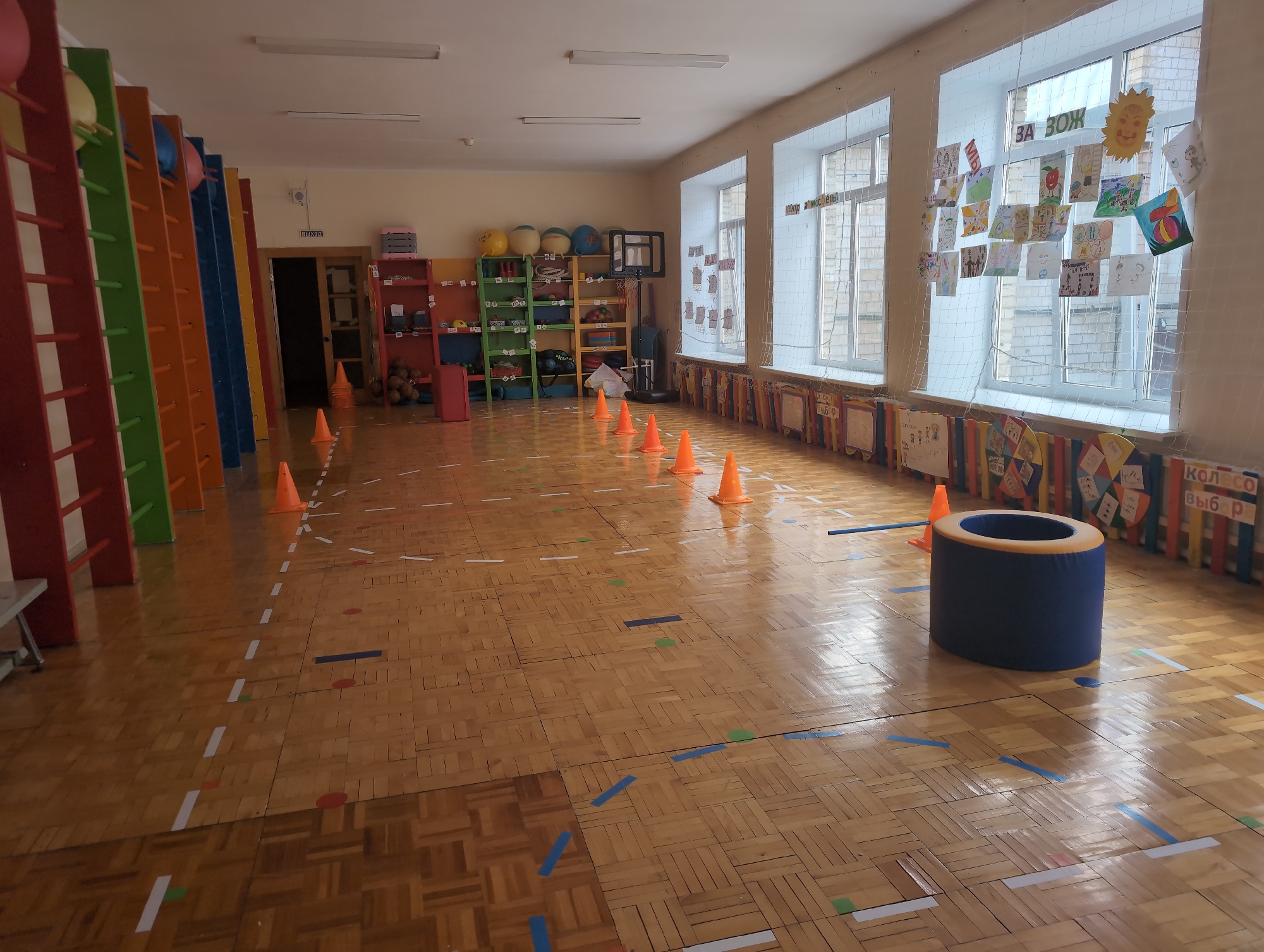 This image depicts a brightly lit indoor physical activity or play area, characteristic of a school gymnasium or a kindergarten sports hall, located in Izhevsk, Russia. No people are present in the scene.
The room features a highly polished wooden parquet floor, extensively marked with white dashed lines and solid colored tapes (blue, green, red), suggesting defined pathways or stations for games and exercises. A series of orange traffic cones are arranged in a line down the center of the floor, indicating an obstacle course or a designated path. In the right foreground, a large, dark blue soft cylinder with an orange rim stands upright, possibly used as a target, seating, or climbing aid.
Along the left wall, several colorful wooden climbing frames or ladders (red, green, orange, blue) are positioned. Further back, a red door is visible, above which the Russian word "ВЫХОД" (Exit) is posted. The back wall holds various shelving units filled with educational toys, balls, and sports equipment, including a small freestanding basketball hoop.
The right wall features large windows, covered with a white safety net from which numerous children's drawings are displayed. These artworks include vibrant colors and various subjects, with a prominent yellow smiling sun. Below the windows, a row of colorful decorative panels further enhances the playful atmosphere.
Additional Russian texts are visible: "ЗА ЗОЖ" (For a Healthy Lifestyle) and "ЦЕНТР ЗДОРОВЬЯ" (Health Center) are displayed on the windows among the drawings. On one of the decorative panels below the windows, "КОЛЕСО ВЫБОР" (Wheel of Choice) is written. The bright natural light filtering through the windows suggests it is daytime.