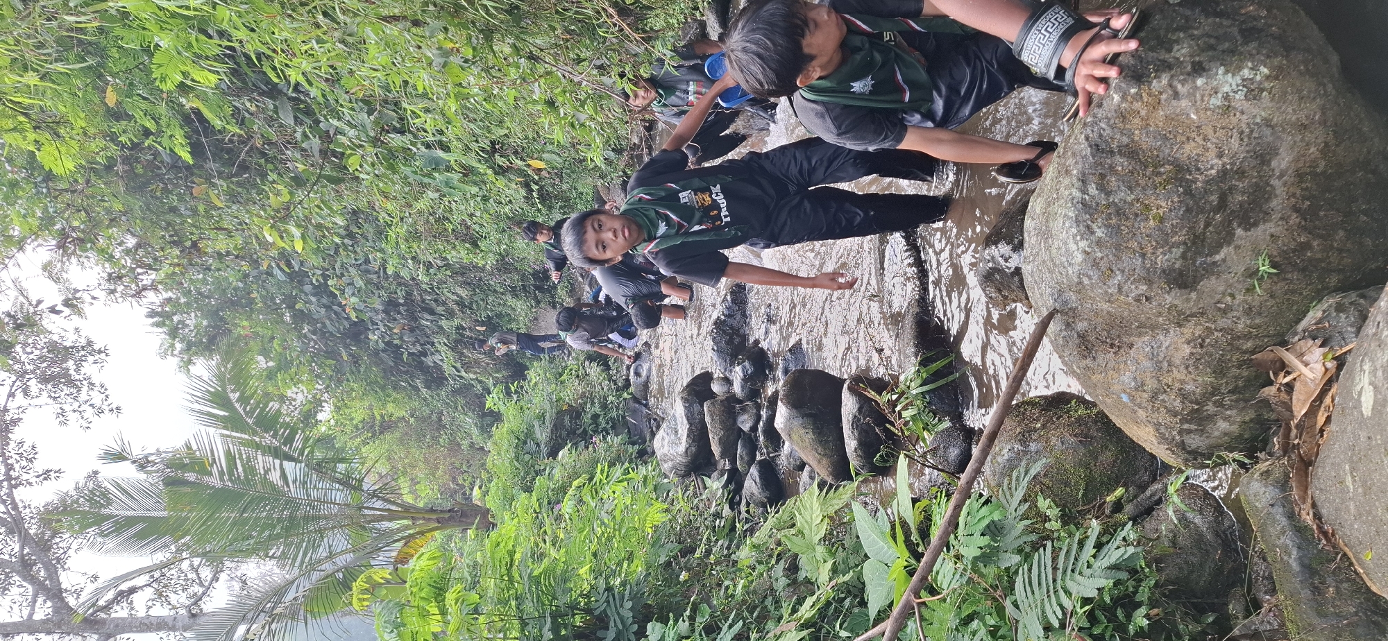 In the city of Puntukrejo, Indonesia, a group of young Scouts navigates a shallow, rocky stream. The water is murky brown, reflecting the overcast sky and the dense, verdant foliage that crowds the stream banks. Towering trees and lush green plants create a canopy overhead, dappling the scene with muted light. The Scouts, clad in dark t-shirts and black pants, are mostly submerged up to their knees or thighs in the water. Several wear green neckerchiefs, indicating their affiliation. They are in various stages of traversing the stream, with some actively walking and others bending to navigate the slippery rocks. One Scout in the foreground looks directly at the camera, his expression serious. Another Scout behind him holds a blue plastic object. The stream bed is littered with smooth, grey river stones of varying sizes. The overall atmosphere is one of outdoor adventure and exploration in a natural, humid environment.