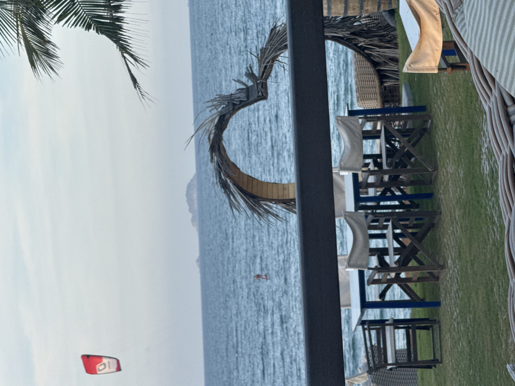 This outdoor scene, possibly in Đà Nẵng, Vietnam, depicts a coastal setting on an overcast day. The focus is on a beachside restaurant or resort area. The scene includes tables and chairs arranged on a patch of grass, suggesting an outdoor dining or relaxation space. A decorative circular structure made of dried palm fronds is visible in the background, partially framing the view of the ocean. A person is kitesurfing. In the distance, there appears to be a mountain or land formation. Palm leaves partially obscure the upper-left corner of the frame, adding to the tropical ambiance. The scene also includes kites in the distance.