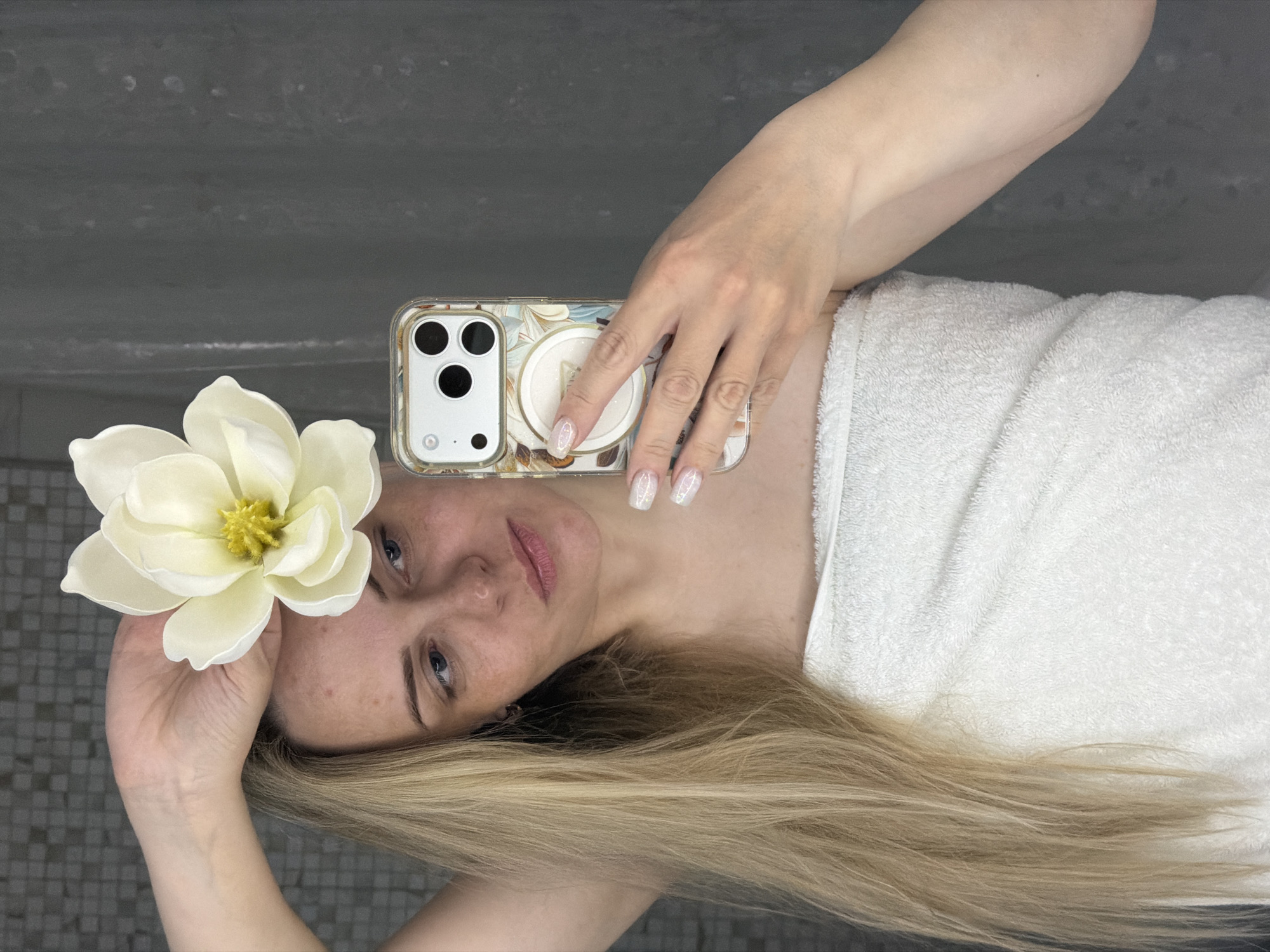 An overhead shot shows a woman lying down, angled horizontally across the frame, with her head towards the left and body extending to the right. She has long, straight blonde hair fanned out behind her and is wrapped in a fluffy white towel. Her left arm is raised, holding a large, artificial white flower with a yellow center near her head. Her right arm is bent, holding a smartphone with a clear, decorative case featuring a floral or abstract pattern and a white pop socket. Her nails are manicured with a light, possibly iridescent, polish.
The woman has a pale complexion, light blue eyes, and visible freckles or blemishes on her face. Her lips are naturally pink, and her expression is neutral and calm, looking directly towards the camera. The background appears to be a grey tiled surface, possibly a wall or floor, with varying shades and some lighter marks, suggesting an indoor setting like a bathroom or spa. The location context is Votkinsk, Russia. There is no visible text.
