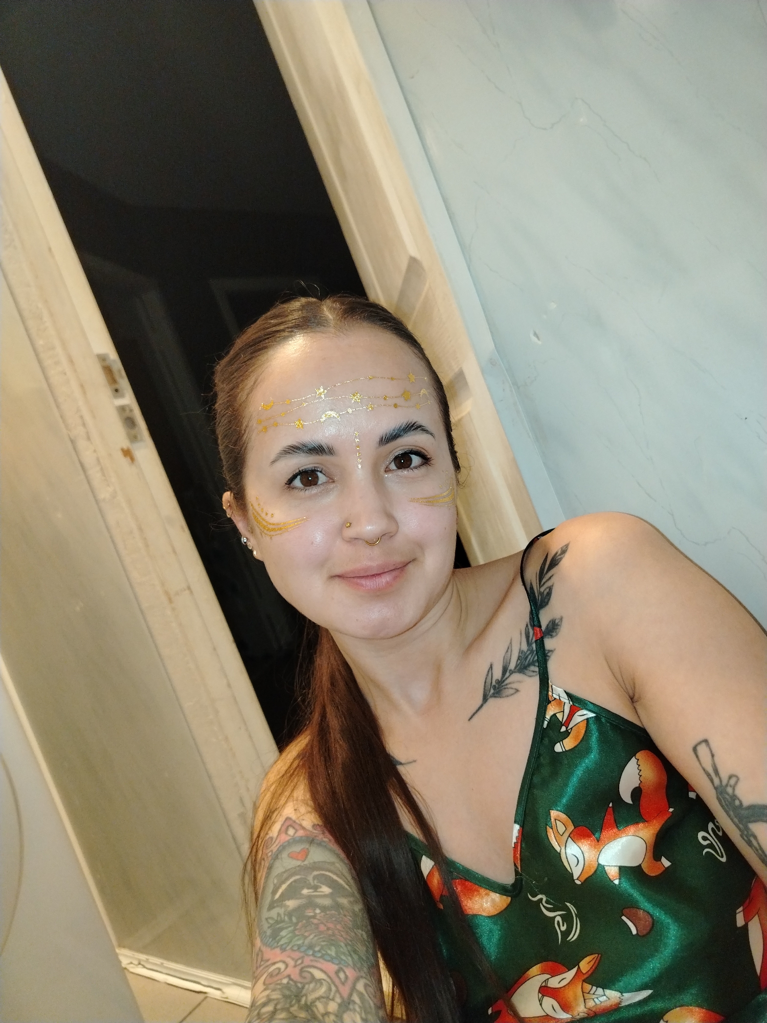 A medium shot captures a woman taking a selfie indoors. She has long, dark brown hair pulled back, and her face is adorned with golden temporary tattoos, featuring stars, moons, and decorative lines on her forehead, and swirls on her cheeks. She looks directly at the camera with a gentle smile. Her brown eyes are prominent, and she has a gold stud piercing in her left nostril and a gold hoop in her right.
She wears a dark green, satin-like camisole or top with a distinct pattern of sleeping and active orange and white foxes, along with some abstract white shapes. Visible tattoos include a delicate leafy branch on her left collarbone, a detailed colorful tattoo on her left upper arm featuring a raccoon head and intricate patterns, and another tattoo on her right upper arm. The background on the left appears to be a dark open space, possibly a doorway, while the right side shows a light-colored wall with faint grey marbling. The lighting suggests an indoor setting, likely with artificial illumination. No text is visible.