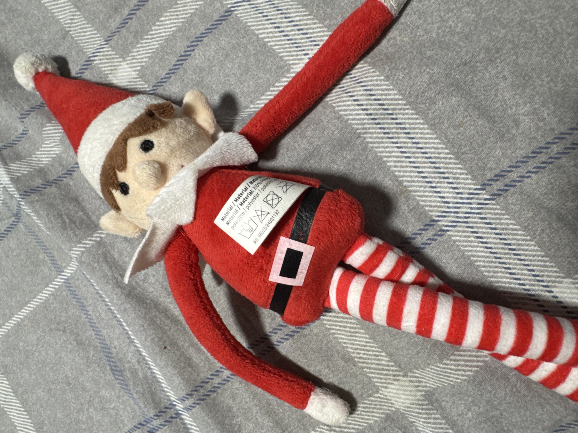 A plush elf doll with a red Santa hat, red suit, black belt with a pink buckle, and red and white striped legs is lying on a grey, plaid blanket. The elf has brown hair and black eyes. The blanket has a plaid pattern of grey, white, and blue lines. There is a tag attached to the elf's waist with writing and symbols. The scene is indoors and appears to be a close-up shot of the doll.