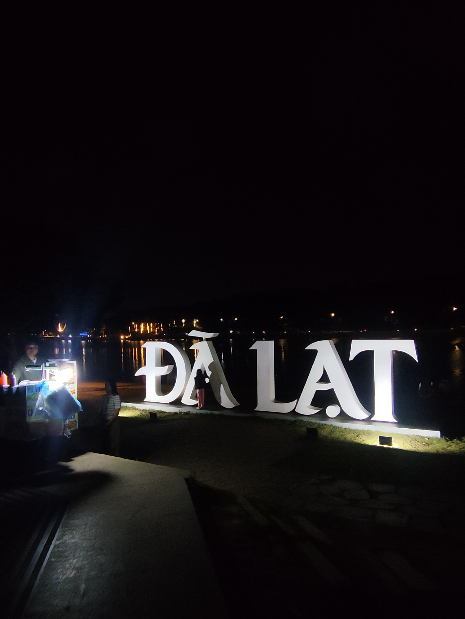 At night in Da Lat, Vietnam, a large, illuminated "ĐÀ LẠT" sign stands on a grassy area near a body of water. A person is positioned near the "A" of the sign. A food vendor cart is visible to the left, attended by a person. Another person is standing nearby. Lights are visible in the distance across the water. A stone walkway is in the foreground.