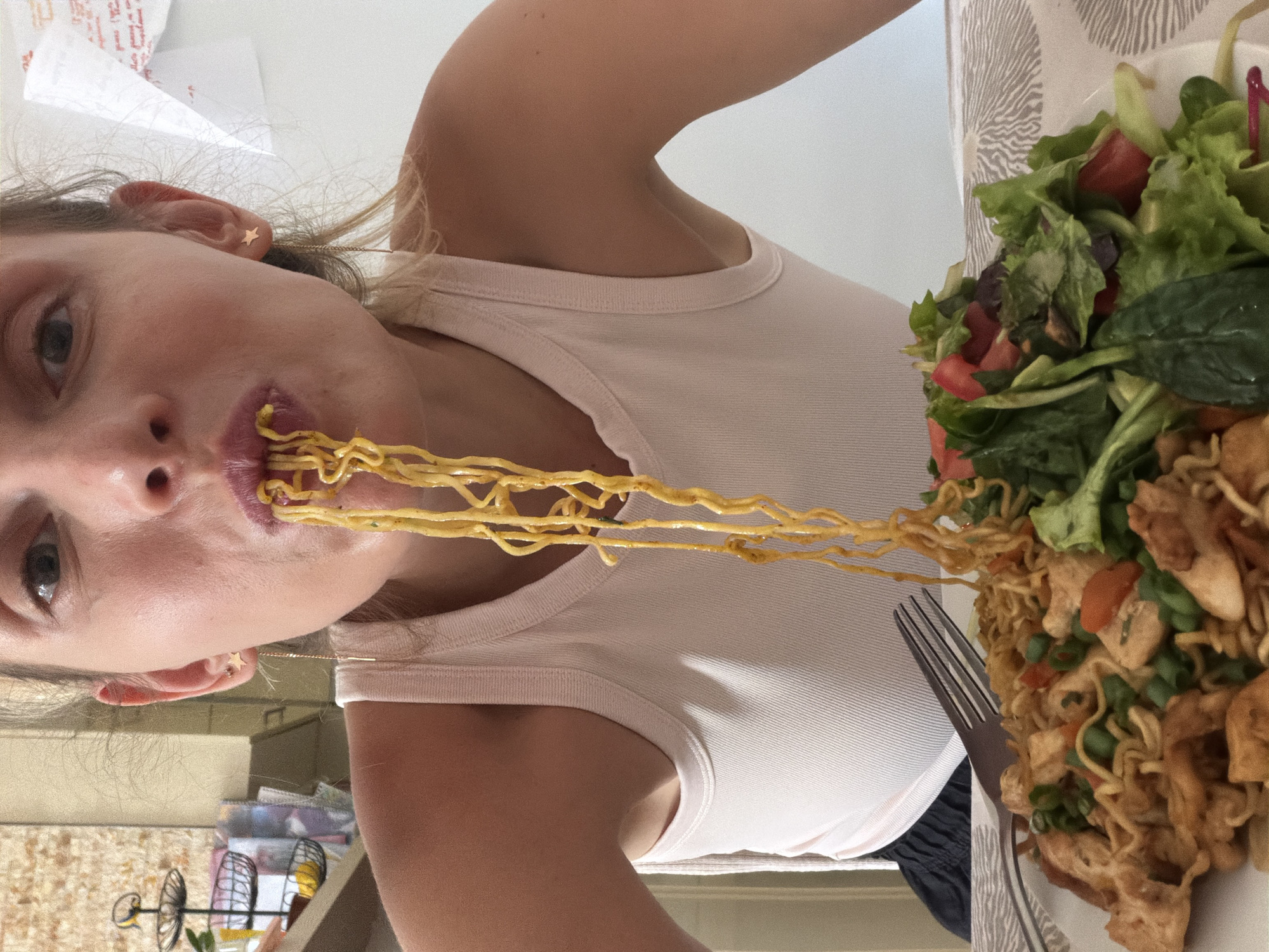 A close-up shot shows a woman eating noodles in Budva, Montenegro. The woman is wearing a white tank top and has star-shaped earrings. Noodles are hanging from her mouth, and a plate of food, including noodles, chicken, and salad, is in front of her. A fork is also visible on the plate. The background is blurred, but it appears to be an indoor setting with a wall and some shelves.