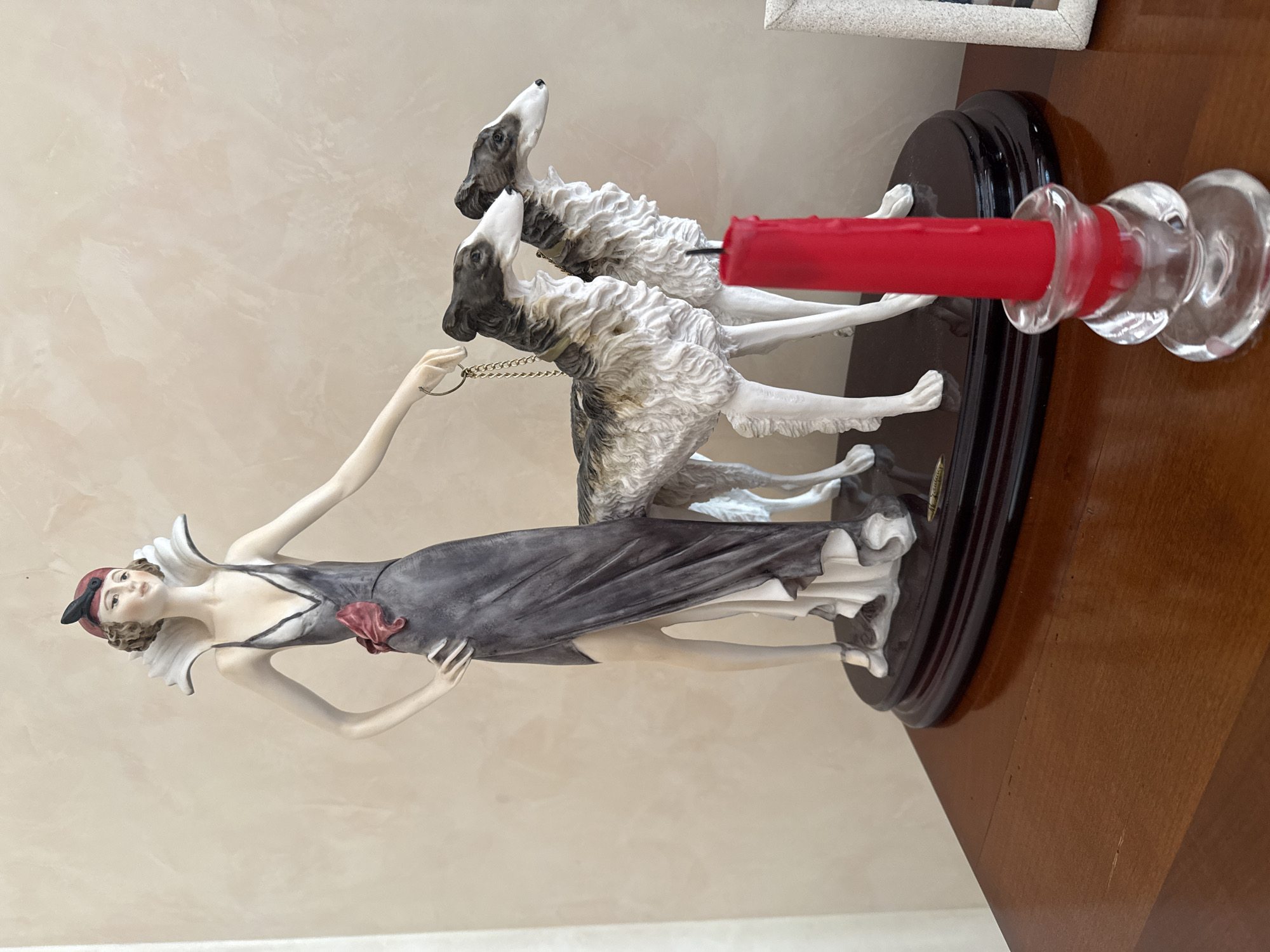 A decorative figurine depicting a woman in an elegant grey dress and a feathered headband, holding the leashes of two borzoi dogs, stands on a polished dark wooden base. The figurine is positioned on a polished wooden surface, possibly a table or dresser. In the foreground, a clear glass candlestick holds a bright red candle. The background is a textured, light-colored wall. The scene is indoors, with the lighting suggesting it is daytime. The location of Yukki, Russia is not depicted or implied by any visual elements in the image.