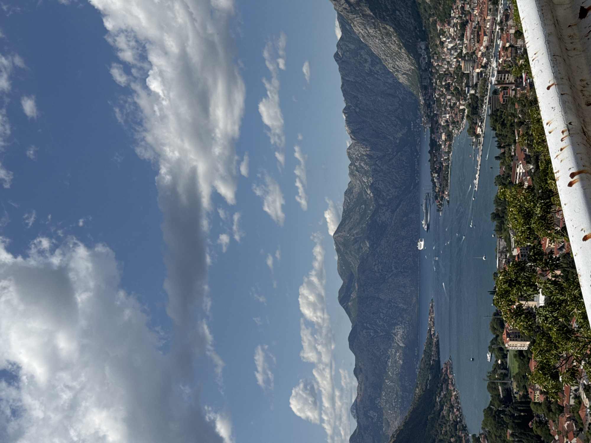 This is a vertical, eye-level, outdoor, daytime photograph taken from an elevated vantage point overlooking the Bay of Kotor in Škaljari, Montenegro. The scene is dominated by a large body of blue water, likely the bay itself, dotted with various vessels including cruise ships and smaller boats. The water is surrounded by steep, rocky mountains that form a dramatic backdrop. On the shoreline, a dense urban settlement with red-tiled roofs and buildings is visible, interspersed with lush green trees. The sky above is a vibrant blue, filled with scattered white and grey clouds. In the foreground, a rusty white metal railing runs horizontally across the bottom of the frame, suggesting the viewpoint is from a road or overlook. The overall impression is a picturesque coastal landscape with a mix of natural beauty and human development.