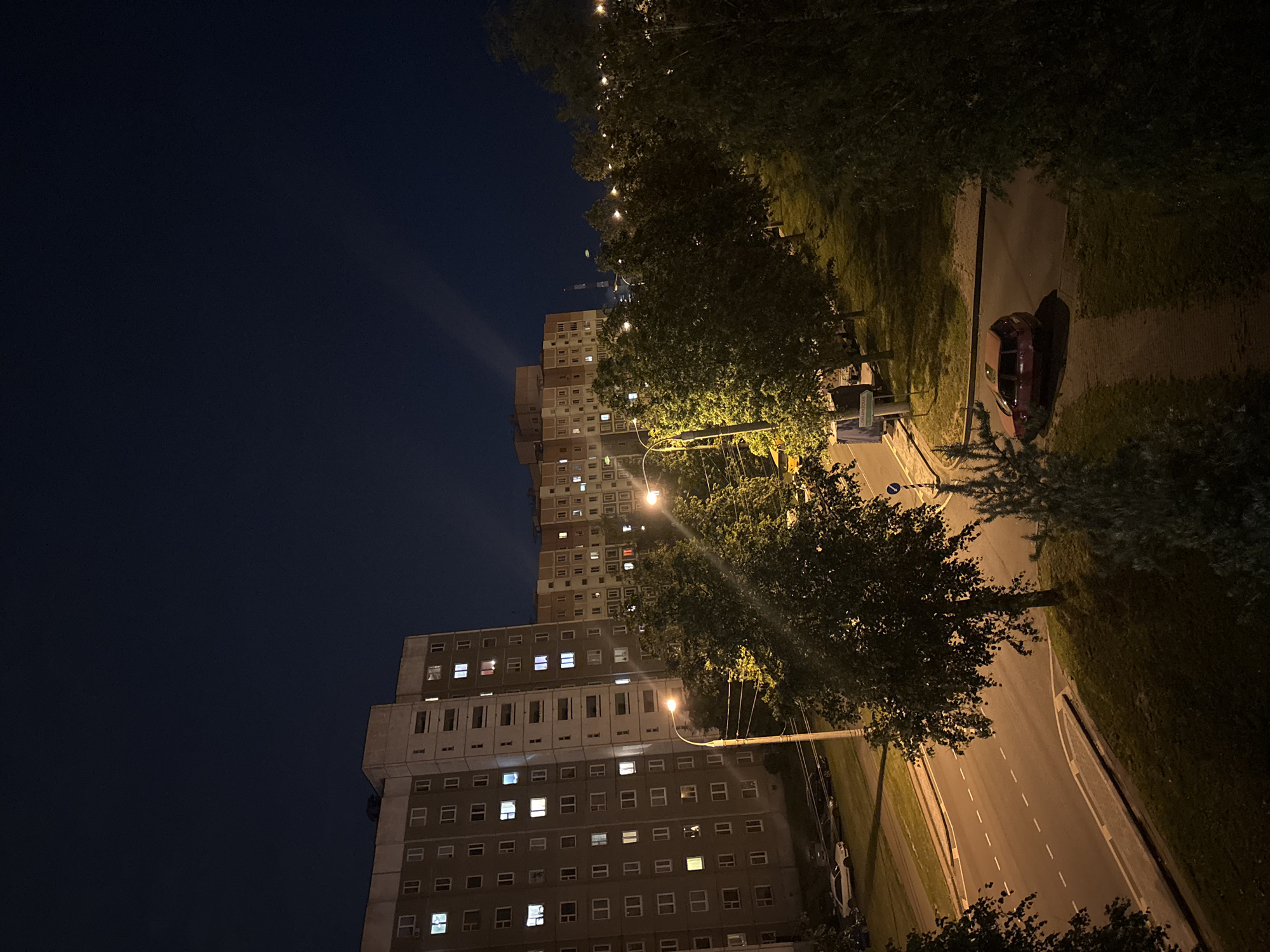 An elevated, full-shot photograph captures a nighttime urban scene in the city of Unknown, Unknown. In the background, several multi-story apartment buildings are visible, their windows illuminated, suggesting occupancy. The sky is dark, with faint light beams visible in the distance. In the midground, trees line a sidewalk and a road, with streetlights casting an orange glow. A red car is parked on a path near the sidewalk. In the foreground, more trees and grass create a shadowed area. The road in the midground shows dashed white lines, indicating traffic lanes. Several parked cars are visible along the roadside near the apartment buildings.