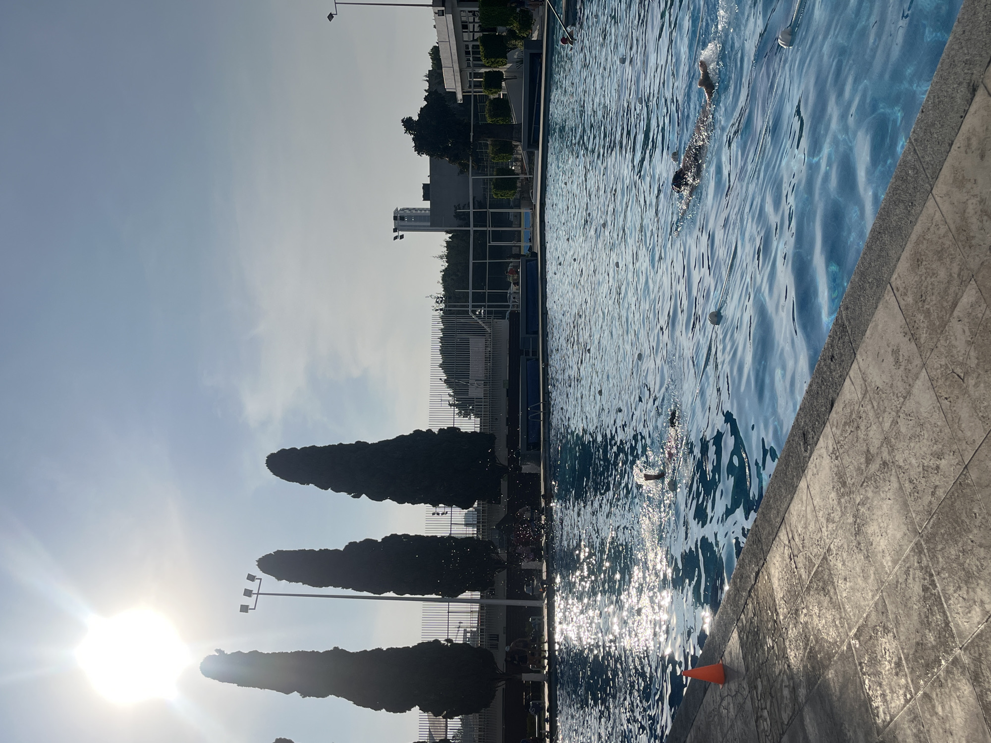 Here's a detailed description of the image:
The image shows an outdoor swimming pool in a city environment. The sky is visible in the upper portion of the image, with the sun shining brightly. Along the side of the pool are tall, narrow trees and lampposts. A fence is partially visible between the trees and the pool area. The water in the pool is blue and reflects the sunlight. People are swimming in the pool. A bright orange cone sits alongside the edge of the pool. The pool's edge has a tiled surface. Buildings are visible in the background, suggesting an urban or suburban setting.