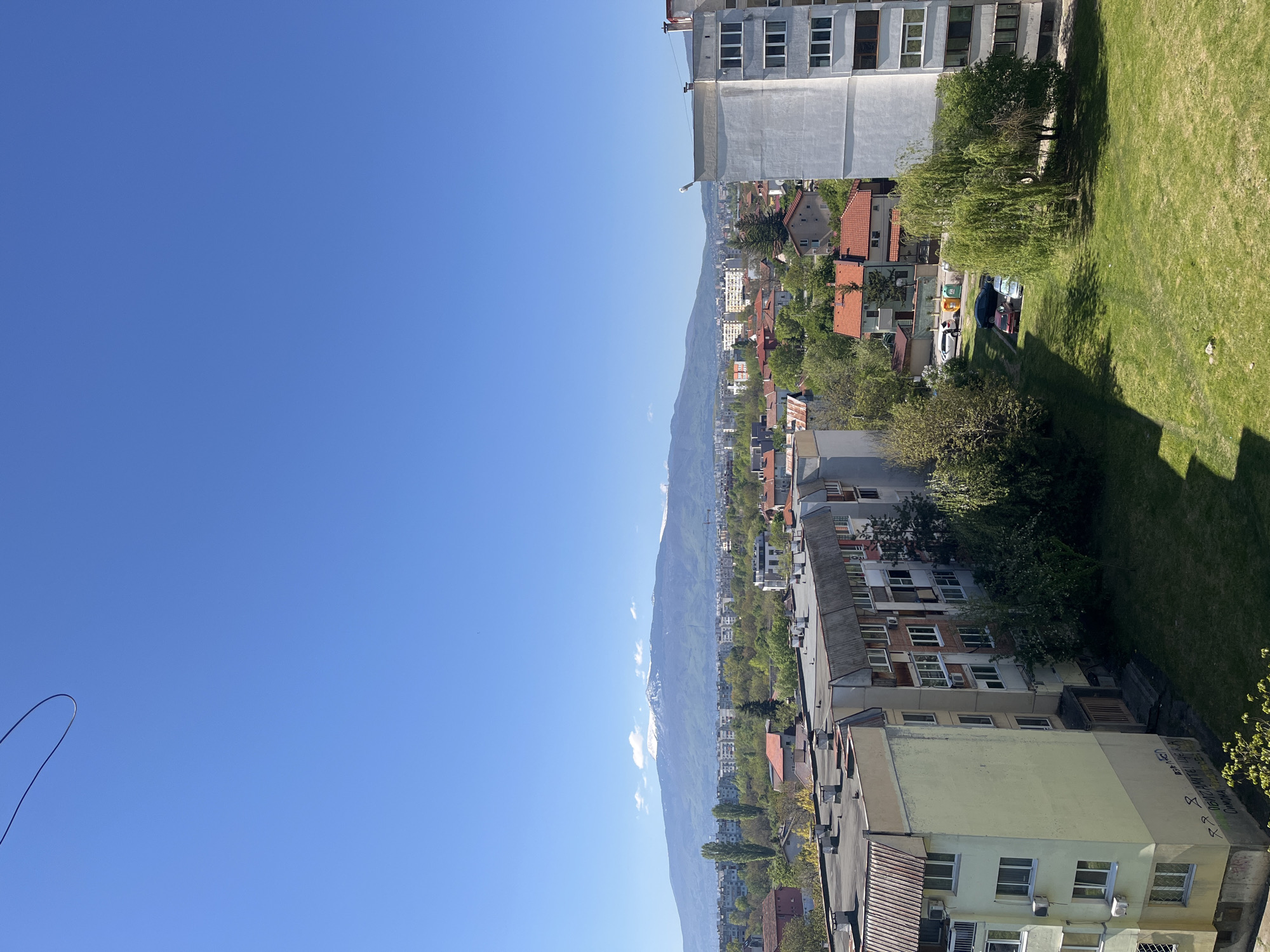 The image shows a view from a high vantage point, likely a tall building or apartment, in Sofia, Bulgaria. The sky is clear and blue. A mountain range is visible in the distance, possibly the Vitosha mountain range. Below, a densely populated urban area is depicted, with a mix of residential buildings, houses with red tile roofs, and green spaces like gardens and parks. There are cars parked along the streets. The foreground shows part of the building from which the photo was taken, including a grassy area and the side of an adjacent building with visible windows and some writing on it.