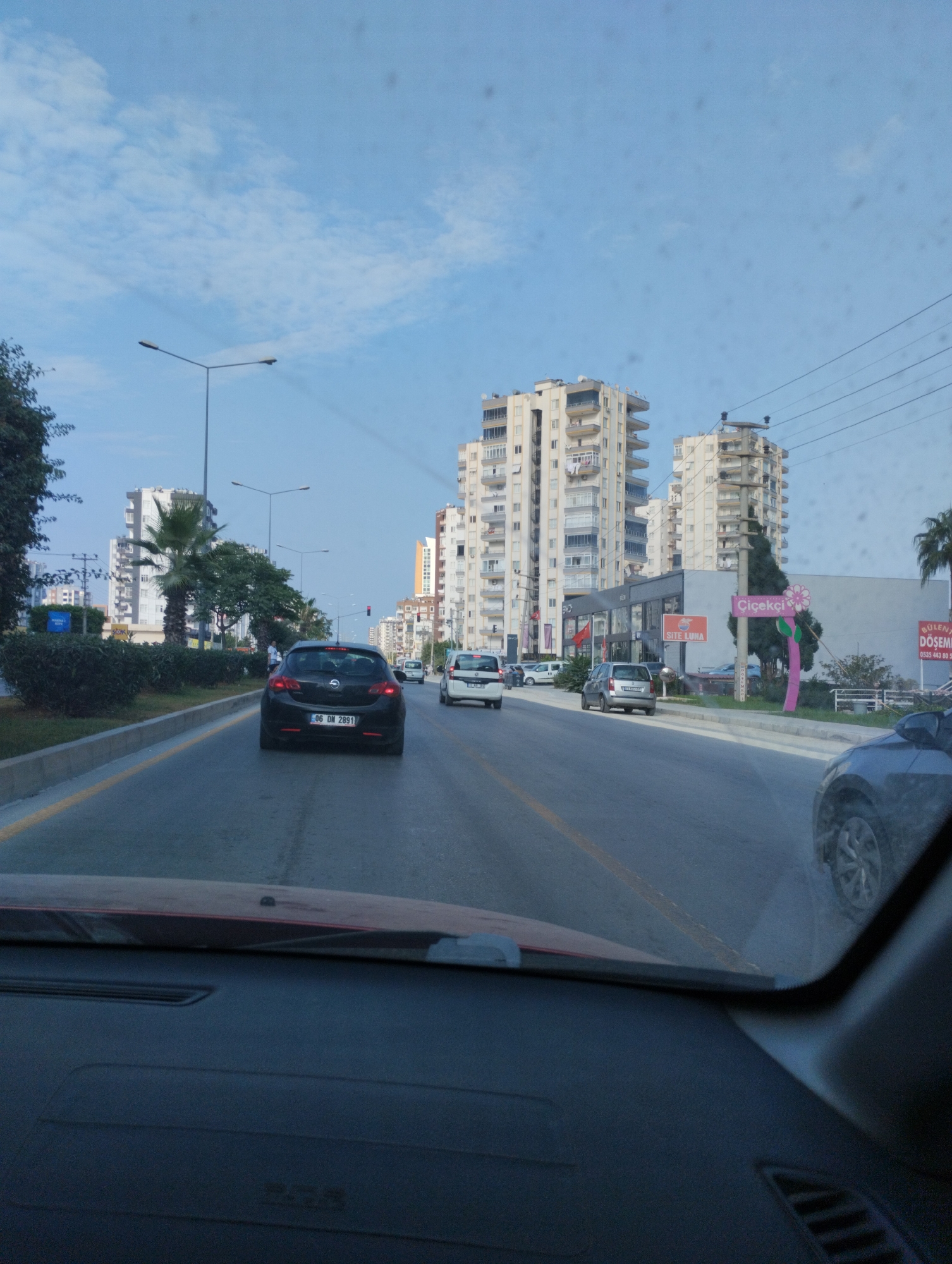The image is a view from inside a vehicle looking forward on a street in Mezitli, Turkey. The street is lined with multistory buildings, some of which appear to be apartment complexes. There are cars on the road, including a black Opel in the foreground with the license plate "06 DN 2891". Streetlights are visible along the road, and power lines cross the sky. The sky is blue with some scattered clouds. A sign with the word "Çiçekçi" (which means florist in Turkish) is visible on the right side of the image, with a pink flower design. The general atmosphere suggests a daytime scene on a city street.