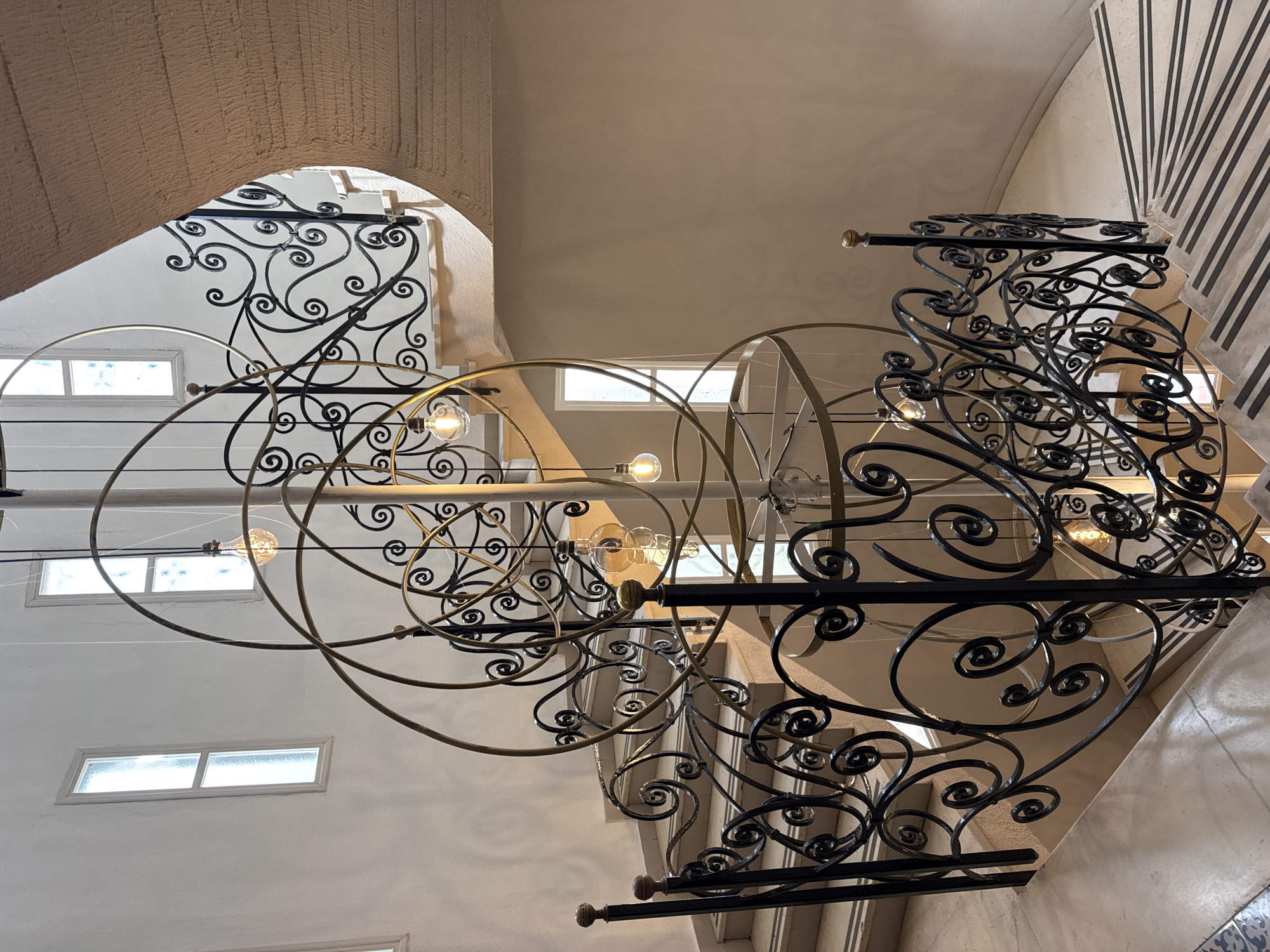 This image captures an upward view within a multi-story interior space, likely a stairwell or atrium, notable for its intricate architectural details. The central subjects are a magnificent, ornate chandelier and an equally elaborate wrought iron balustrade.
The chandelier, suspended prominently, features multiple concentric and intersecting rings or spherical elements made of a brass or gold-toned metal. Numerous exposed filament light bulbs, characteristic of a vintage or industrial aesthetic, are strategically placed along the fixture, casting a warm glow.
The balustrade, made of dark, possibly black, wrought iron, displays an array of flowing scroll patterns and decorative flourishes, indicative of skilled craftsmanship. This balustrade runs alongside a staircase, whose light-colored steps are partially visible.
The surrounding architecture includes light-colored, possibly beige or off-white, walls. Several arched openings, some with a textured, stucco-like finish, are visible, adding to the building's classic feel. Small, rectangular windows with frosted or obscure glass panes are set into the walls, allowing natural light to filter in. In the lower right corner, a small section of decorative blue and white patterned tiles can be seen, hinting at traditional Italian design.
No people are present, and no specific activity is taking place. The scene conveys a sense of elegant, timeless architecture, possibly in a historic building. The combination of natural light from the windows and the artificial illumination from the chandelier suggests either daytime or a brightly lit interior at any hour. No visible text is present. Given the location context of Isola delle Femmine, Italy, the aesthetic aligns with Mediterranean or Italian architectural styles.