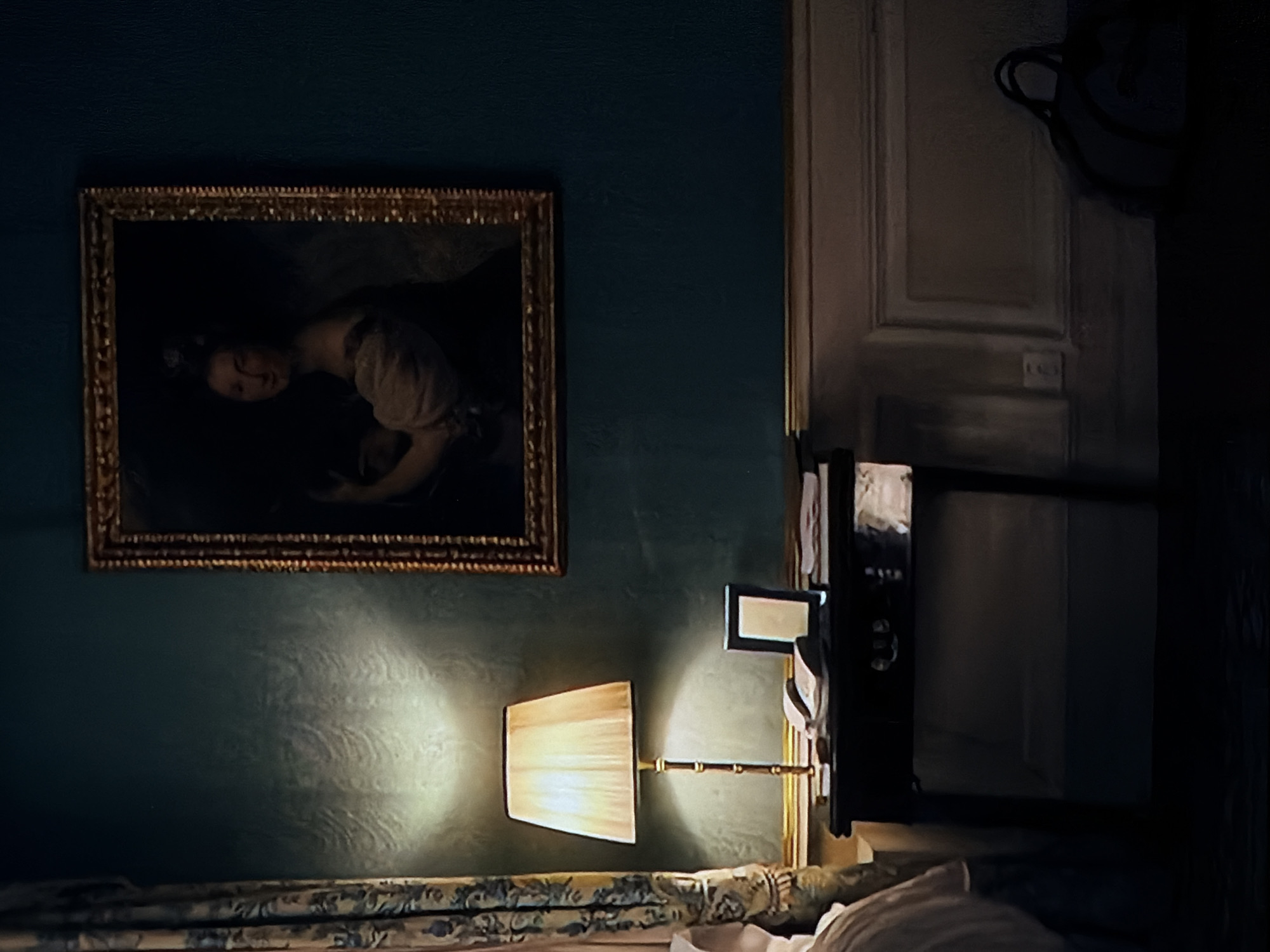 This image presents a dimly lit interior scene, likely a bedroom or lounge area, characterized by a refined and traditional aesthetic. The dominant feature is a dark teal or greenish-blue wall that displays a subtle, swirling damask-like pattern, particularly visible where illuminated.
A rectangular painting, set in an elaborate golden frame, hangs on the wall. The artwork depicts a portrait of a woman with dark hair, seemingly reclining or positioned horizontally, looking downwards. Her light-colored attire suggests a historical or classical style.
Below the painting, a wall-mounted lamp with a light-colored, possibly fabric, shade provides the primary illumination. The lamp emits a warm, soft glow, creating a distinct circular highlight on the textured wall. Its arm appears to be crafted from a golden or brass-toned material with a bamboo-like texture.
At the bottom of the frame, a section of furniture, presumably a bed or sofa, is visible, covered with a patterned fabric featuring floral or botanical designs in shades of blue, green, and cream. To the far right, a portion of a white pillow or cushion is also seen.
To the right of the painting and lamp, a tall, light-colored panel door with classic architectural molding is partially visible, featuring a wall-mounted electrical outlet or switch plate. Adjacent to the lamp and door, a dark, rectangular object, possibly a mirror or a dark television screen, is mounted. An indistinct dark object, potentially a hanging bag or decoration, is vaguely visible above the door.
The overall low light levels strongly suggest the scene takes place during the evening or nighttime. The classical decor elements and subdued lighting create a tranquil and somewhat moody ambiance. No explicit text is visible in the image. Given the location context of Sofiivska Borshchahivka, Ukraine, this room may belong to a residence or a heritage-style accommodation.