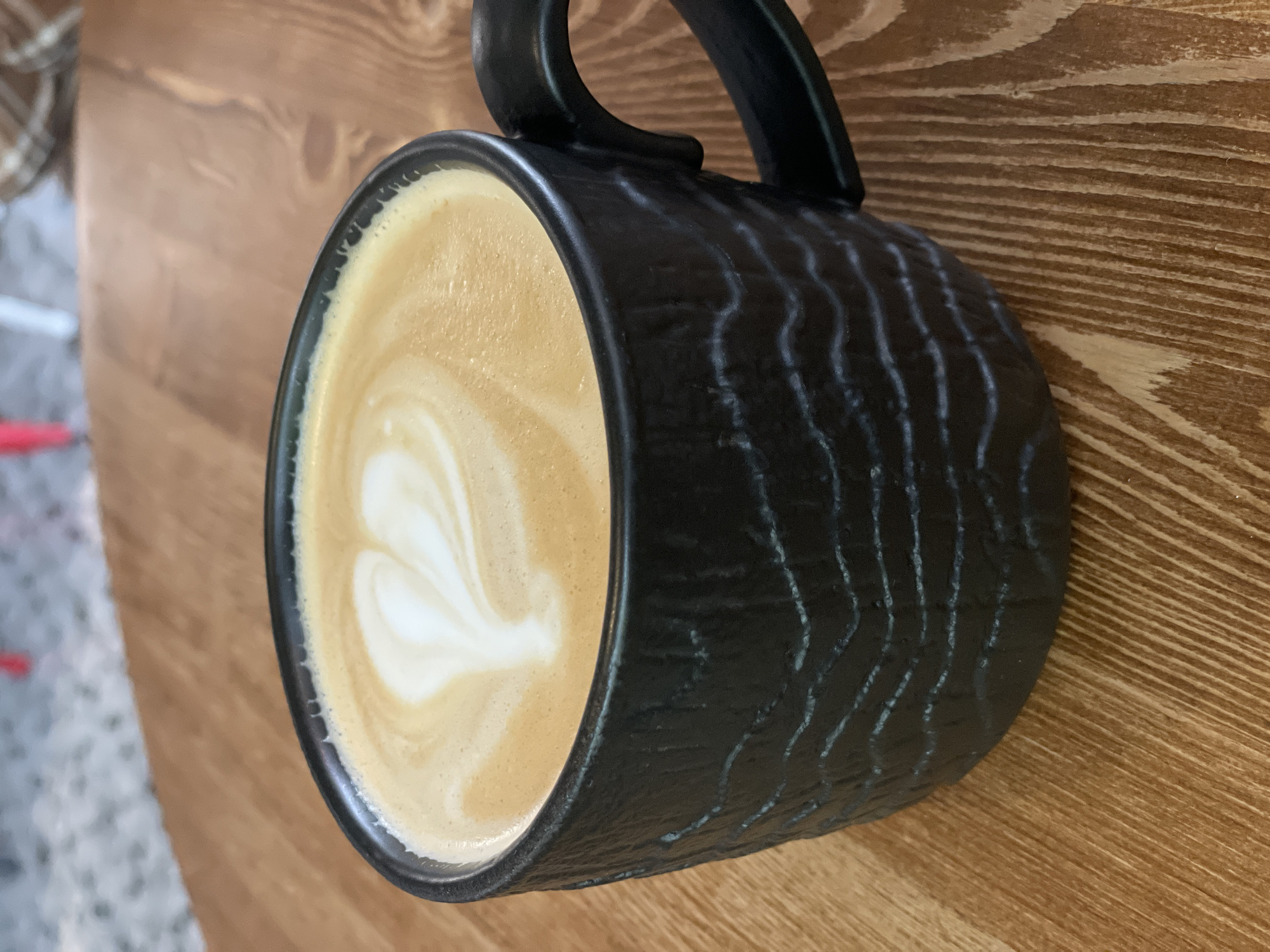 A black textured mug filled with coffee and latte art sits on a wooden table. The latte art forms a heart shape. The background is out of focus and shows a hint of a person and a countertop. The location is not discernible from the image.