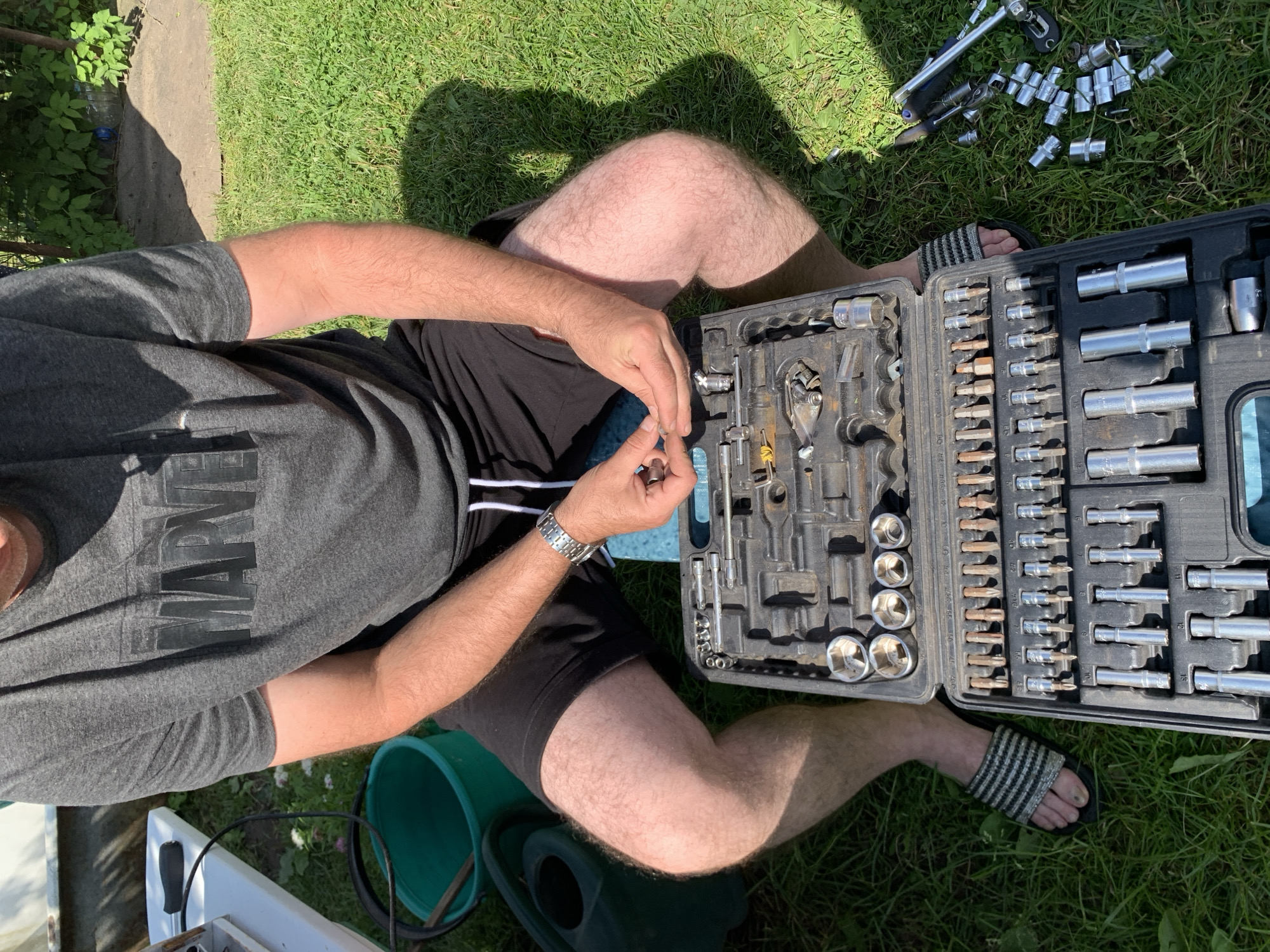 A close-up, high-angle shot shows a person's torso and legs sitting on the grass in an outdoor setting, with a tool kit laid open in front of them. The person is wearing a grey t-shirt with a "MARVEL" logo and black shorts. Their arms are resting on the open tool kit, and their hands are manipulating a small metallic object. The tool kit, a black plastic case, contains an assortment of sockets, bits, and wrenches. To the right of the tool kit, various detached sockets are scattered on the grass. The background shows green grass, some foliage, and a glimpse of what appears to be a paved area and a green bucket. The overall impression is of someone engaged in a task involving tools outdoors. The location specified, Відзаўскі сельскі Савет, Belarus, is an administrative division which suggests a rural or semi-rural environment.