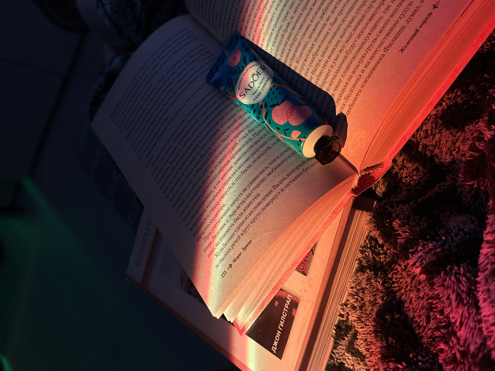 Here is a detailed description of the image:
**Scene:** An open book lies flat on a textured surface, possibly a rug or blanket. A partially used tube of "SADOER Peach & Plants" hand cream rests on the right-hand page of the book. The light source is from the right, creating a warm glow on the book and the surrounding area, while the left side is in relative shadow.
**Objects:**
* **Book:** An open book with text in what appears to be Russian, judging by the Cyrillic alphabet. The book has a red cover. The pages are slightly yellowed. The spine displays the author's name as "ДЖОН ГИЛСТРАП" and the title is "ИЗБРАНИ".
* **Hand Cream Tube:** A partially used tube of hand cream with the brand name "SADOER Peach & Plants." The packaging features a turquoise background with stylized peach and plant designs. The tube has a black cap.
* **Textured Surface:** The book is resting on a soft, textured surface that seems to be a rug or a blanket. The texture is dark, and it catches the light.
**Lighting:**
* The lighting is warm and artificial, likely coming from a lamp or other indoor light source positioned to the right of the frame. The light casts shadows and highlights, enhancing the textures.
**Overall Impression:** The scene has a relaxed, cozy vibe, suggesting someone is enjoying a quiet moment with a book and hand cream in a dimly lit setting. The presence of the hand cream on the open book suggests it is being used while reading.