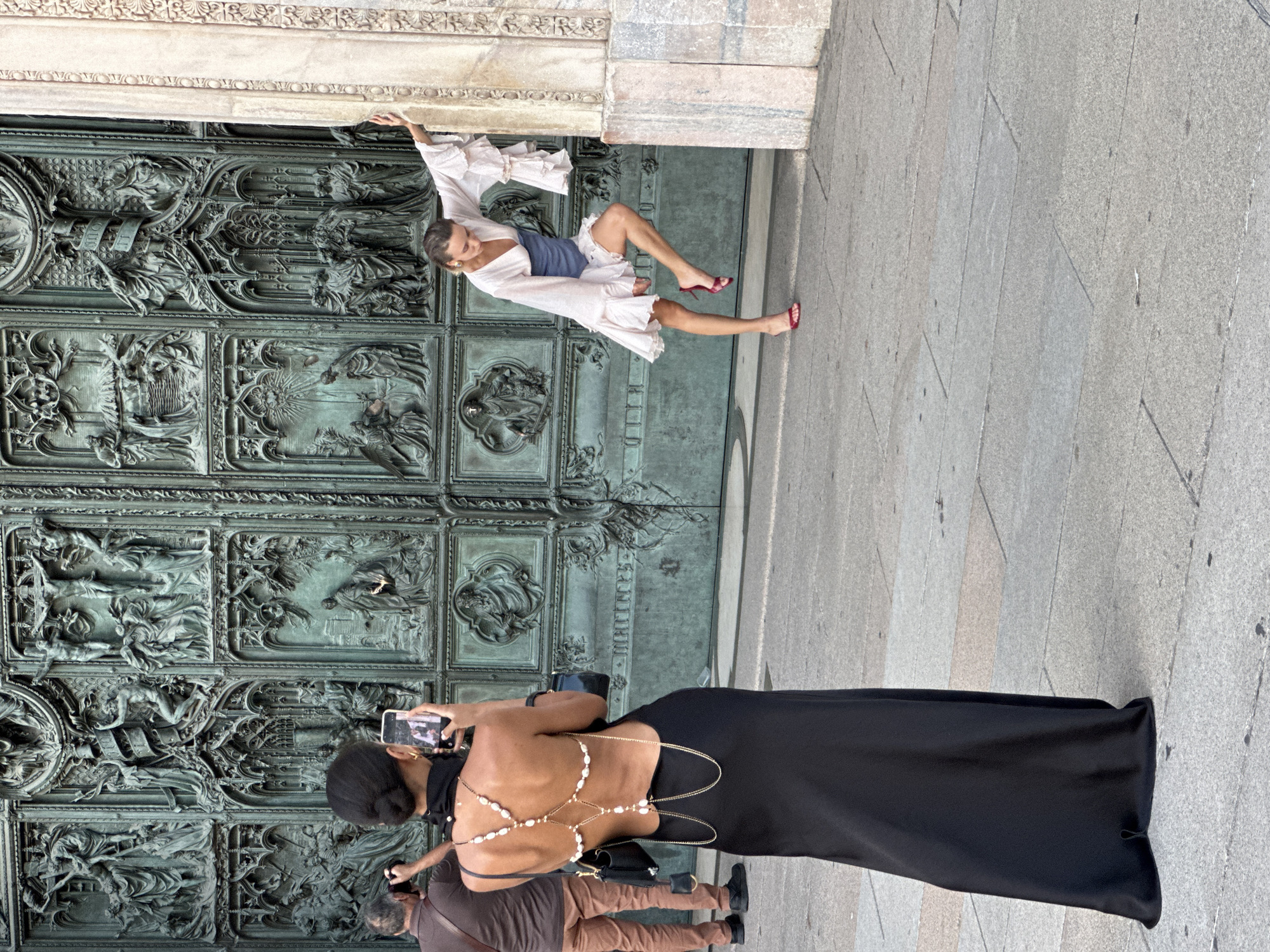 In Milan, Italy, outside the Duomo di Milano, a woman in a black dress with an open back adorned with pearls and chains is taking a photo with her smartphone of another woman. The second woman, in a light-colored ruffled dress and red heels, is posing provocatively against a pillar. In the background, a man with a camera is visible. The Duomo's large, ornate bronze doors serve as the backdrop.