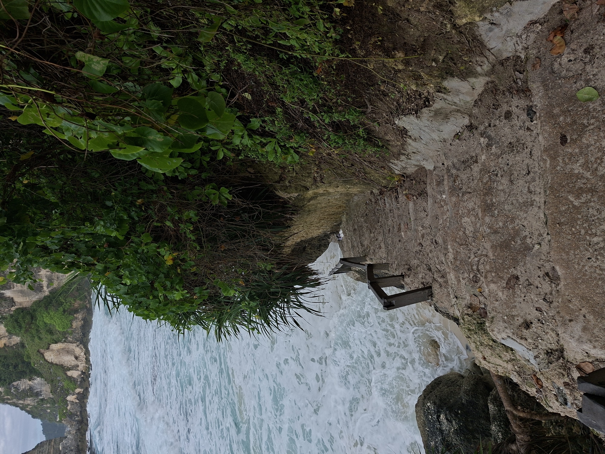 This image depicts a coastal scene in Nusa Penida, Indonesia. A set of concrete stairs leads down from a cliffside covered in vegetation. Waves crash against the base of the cliff and a rocky outcrop near the stairs. A wooden handrail is partially visible along the stairs. The ocean appears turbulent with white foam. The cliff is covered in greenery.