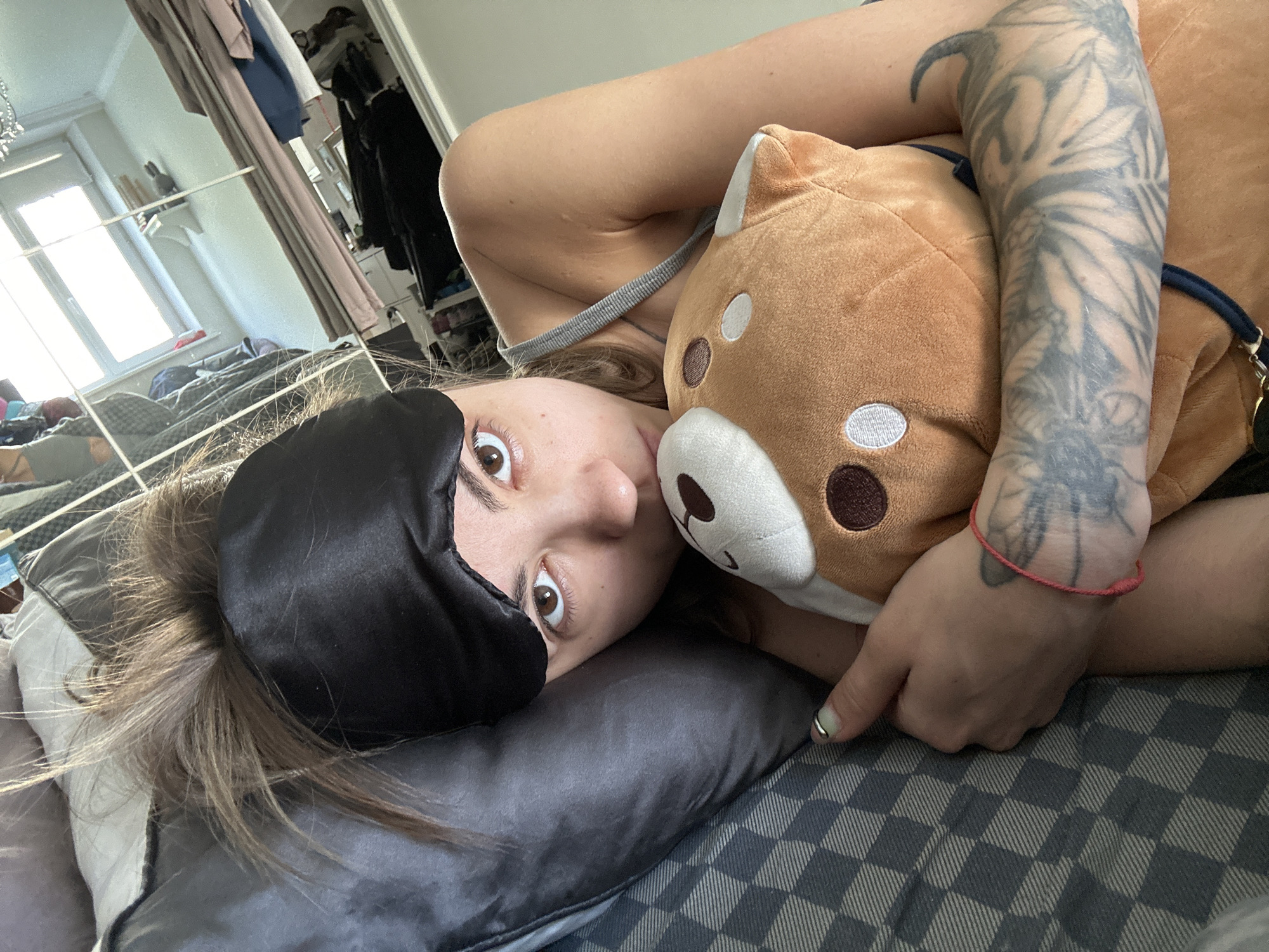 A woman with a black eye mask is lying on a bed, holding a stuffed animal. Her arm is covered in tattoos. The room appears to be a bedroom, with a closet and a window visible in the background. The woman is looking at the camera with wide eyes. The scene is lit by natural light coming from the window.