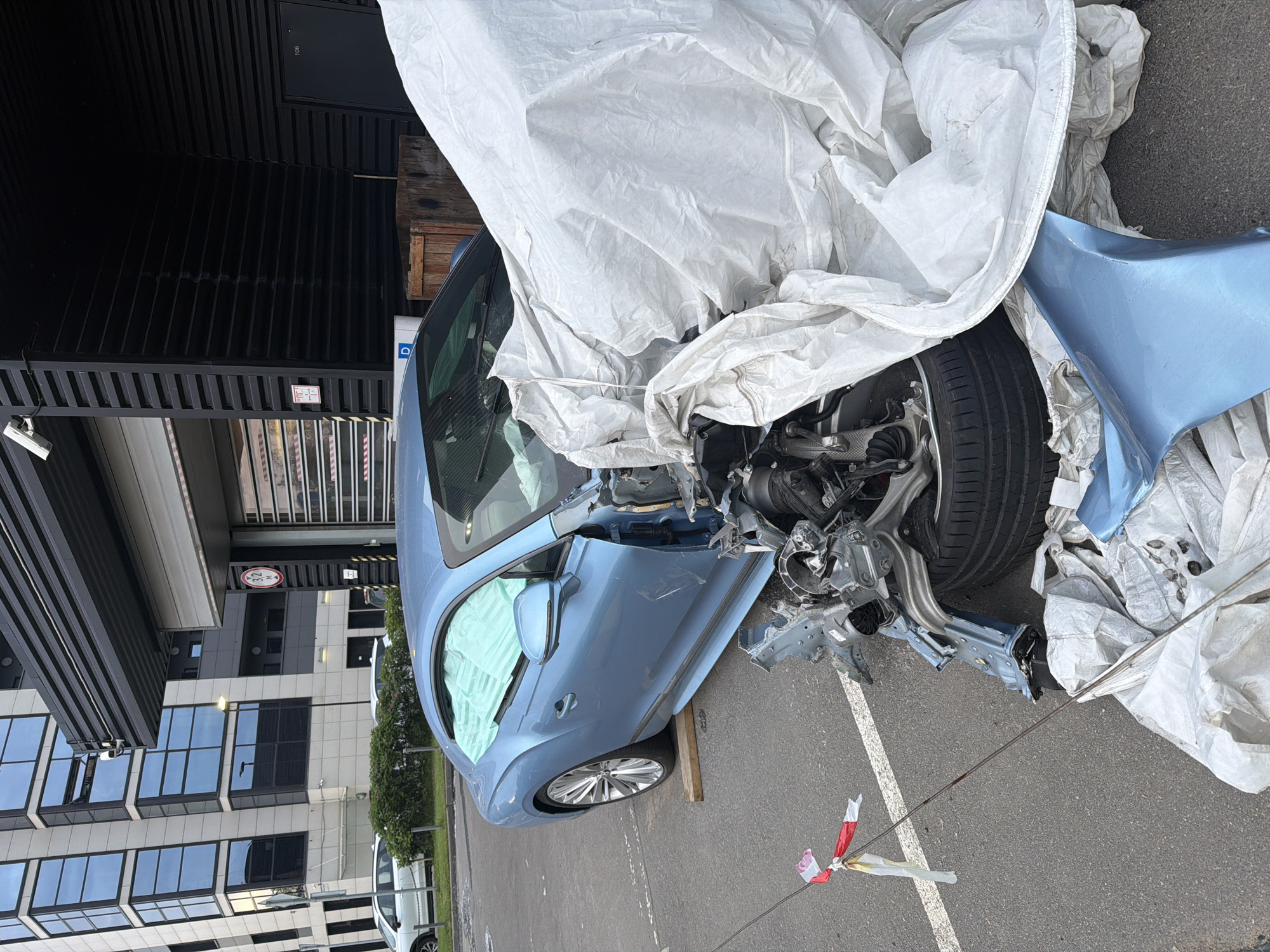 In an outdoor urban setting, a light blue Bentley Continental GT is parked on asphalt. The front passenger side of the car has sustained significant damage, with the bumper, hood, and fender appearing to be missing or heavily compromised. The internal mechanical components of the car, including suspension and engine parts, are exposed. A white tarp is draped over the front of the vehicle, partially obscuring the extent of the damage. The car is supported by wooden blocks under the front passenger-side tire. Red and white tape is tied to a wire that runs across the ground in front of the car. In the background, a modern building with glass windows and a dark, corrugated facade stands behind the car. Another white car is visible further in the background. A street sign with a red circle and a number "3.2" inside is mounted on the building's facade.