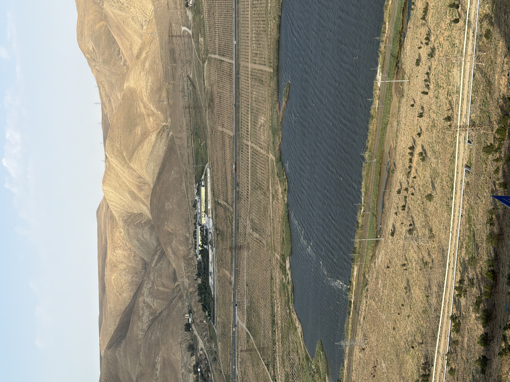An aerial, elevated view captures a dry, arid landscape with undulating brown hills in the background. A body of dark, rippling water occupies the middle ground, with reeds growing along its edge. Adjacent to the water is a paved road with several vehicles, appearing to be cars and trucks, traveling in both directions. Beyond the road, a grid-like pattern of vegetation, possibly agricultural fields or orchards, stretches towards the hills. In the far distance, a cluster of buildings, likely a factory or industrial complex, is visible. Several tall, lattice-like electricity pylons are scattered throughout the scene, indicating infrastructure. The sky is a pale blue with minimal cloud cover. The overall impression is one of a semi-arid environment with evidence of human development, possibly located on the outskirts of Baku, Azerbaijan, given the characteristic landscape and infrastructure often seen in its surrounding areas.