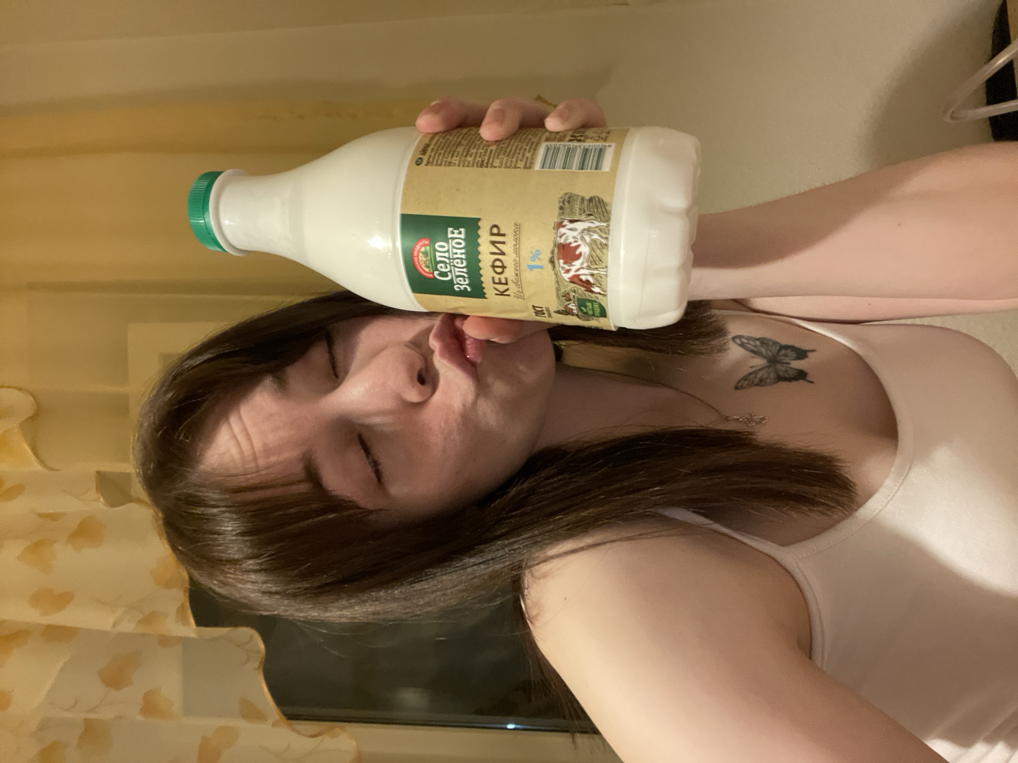 A person with long dark hair is holding a bottle of kefir in their right hand, nearly touching their lips with the bottle. The bottle has a green cap and a label in Russian, which includes the words "Кефир" and "Село Зелёное" (Kefir and Green Village). The label also indicates it's a 1% fat kefir. The person is wearing a white tank top, and a butterfly tattoo is visible on their chest, near their right collarbone. A silver necklace is around their neck. The background shows a patterned curtain and a window. The exact location in the city of Chaikovsky, Russia, is not determinable from the image.