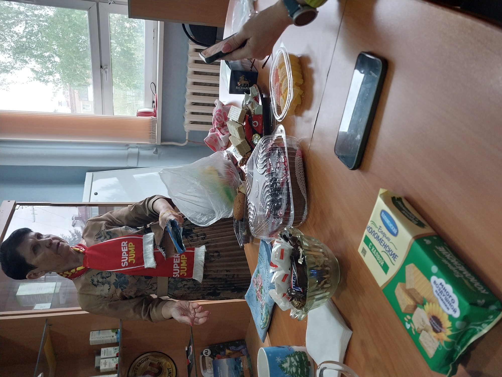 The image shows an indoor scene in Orenburg, Russia. A person, possibly a woman, is standing near a wooden shelf filled with various items. She is wearing a brown patterned shirt, brown skirt, and a red scarf with the words "SUPER JUMP" written on it.
On a wooden table, there are several food items, including cakes, candies in a glass bowl, waffles in a green package labelled "Коломенские", and jellies in a plastic container. Additionally, there is a smartphone, a box of Richard tea, a blue patterned mug, and some napkins on the table.
Another person is visible holding a smartphone above the table, likely taking a photo. A window in the background shows a view of a tree outside. A radiator is also visible near the window.