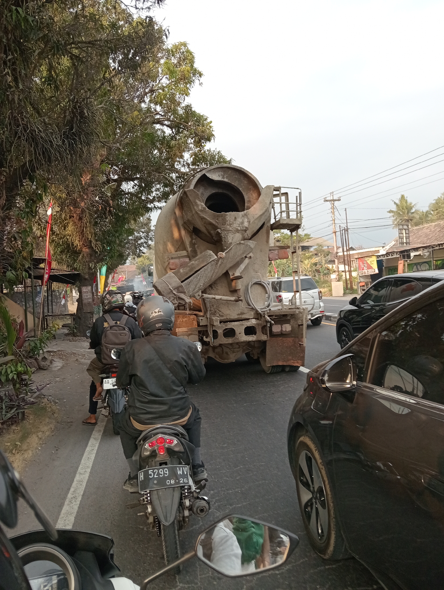The image depicts a traffic jam on a road in Salam, Indonesia. A large cement mixer truck is a dominant subject, positioned in the center of the frame, its drum filled with cement. Several motorcycles are visible, with riders wearing helmets and jackets, navigating the congested road. One motorcycle in the foreground has a license plate that reads "H 5299 WV 08-26". A black car is partially visible on the right side of the image. The environment includes trees lining the road, telephone poles, and buildings in the background, suggesting a semi-urban or suburban setting. The sky is overcast, indicating a potentially cloudy day.