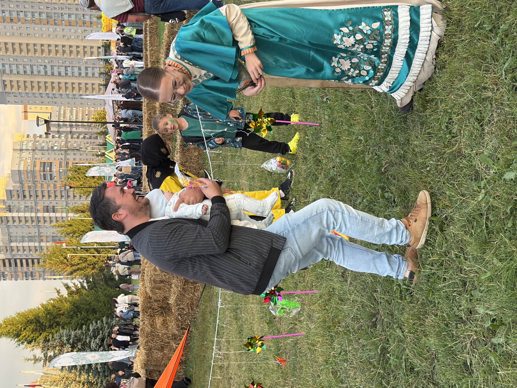 This is a vertical, full-shot, eye-level photograph taken outdoors during the daytime. A man is holding a baby and smiling while looking at the baby. The man is wearing a grey pinstripe jacket, a white shirt, and light blue jeans. The baby is swaddled in a white blanket and wearing a white and pink onesie.
Behind the man and baby, a crowd of people are gathered. Many are standing and talking, some are sitting on hay bales. They appear to be at an outdoor event or festival. In the background, there are tall, modern buildings. Trees are visible as well, and the ground is covered in grass and hay.
To the left of the man, a young girl in a teal dress with ornate gold trim and floral embroidery is visible, looking down at something in her hands. Next to her, a young boy with face paint is looking towards the camera. Several colorful pinwheels are planted in the grass, suggesting a festive atmosphere.
The lighting suggests it is either morning or late afternoon. The overall mood appears happy and relaxed. No legible text is visible in the image.