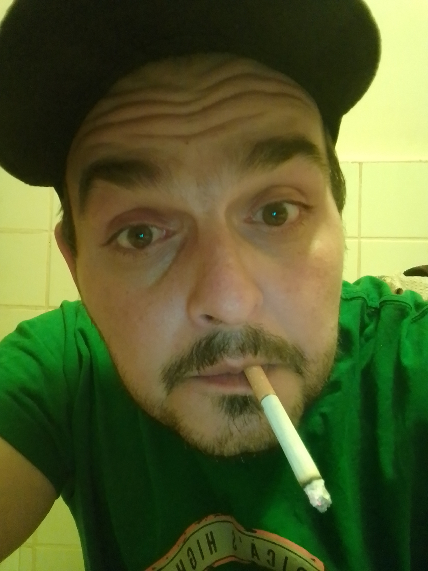 A man wearing a dark hat and a green t-shirt is shown from the chest up, holding a lit cigarette in his mouth. He has wrinkles on his forehead and a mustache. The background appears to be a tiled wall, possibly in a bathroom. The lighting is dim and casts a greenish hue on the scene.