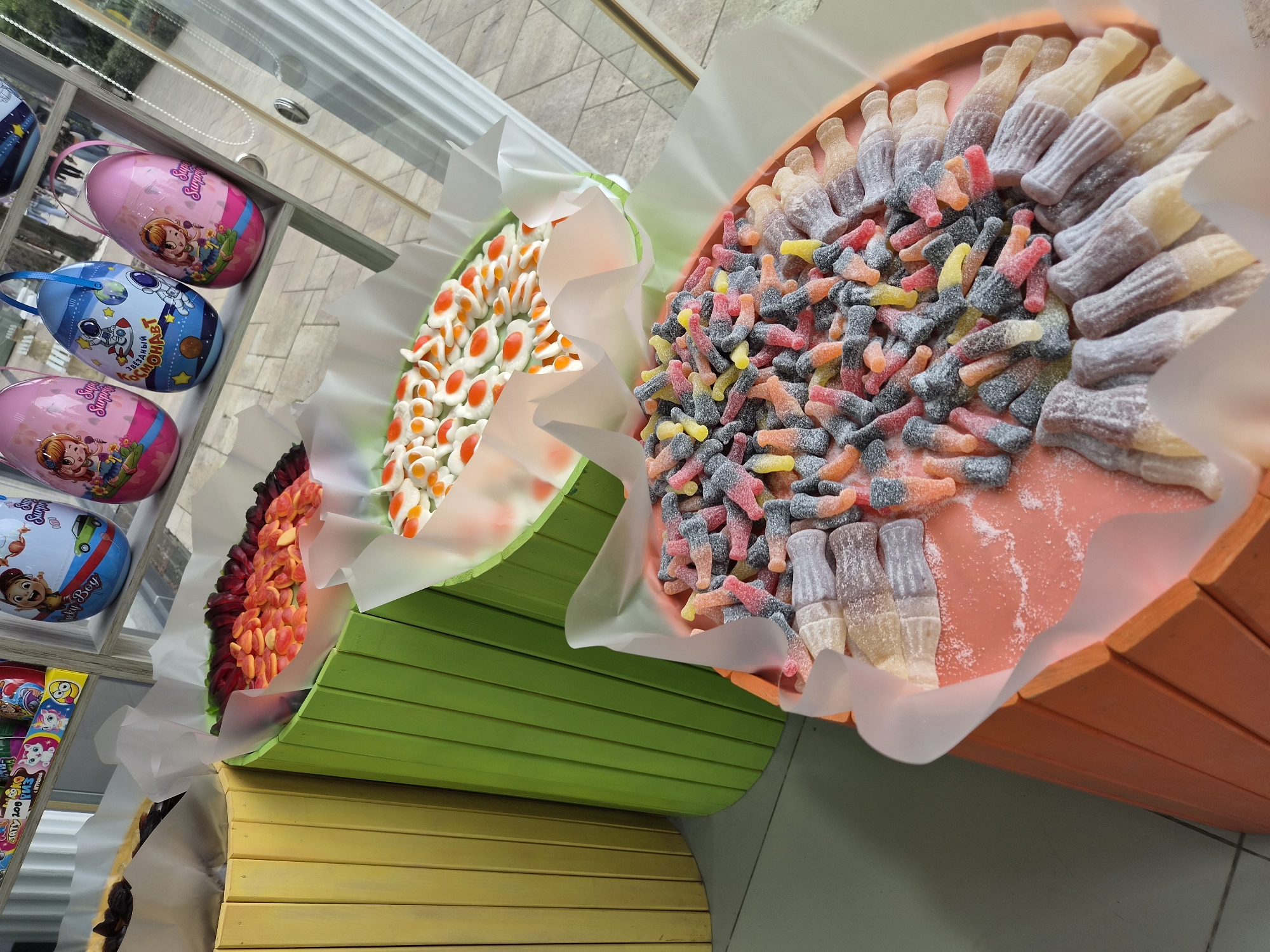 The image captures a display of candy assortments in what appears to be a shop or market setting. There are three colorful, horizontally-ridged containers filled with different kinds of gummy candies. The container colors are red/orange, green, and yellow. The candies include gummy rings, gummy soda bottles, and other varied shapes and colors.
To the left of the candy display is a shelf with egg-shaped containers featuring cartoon characters. The setting appears to be indoors, likely a retail space in Sochi, Russia, given the Cyrillic text on one of the containers. The environment is generally well-lit. The focus is on the colorful and appealing arrangement of sweets for sale.