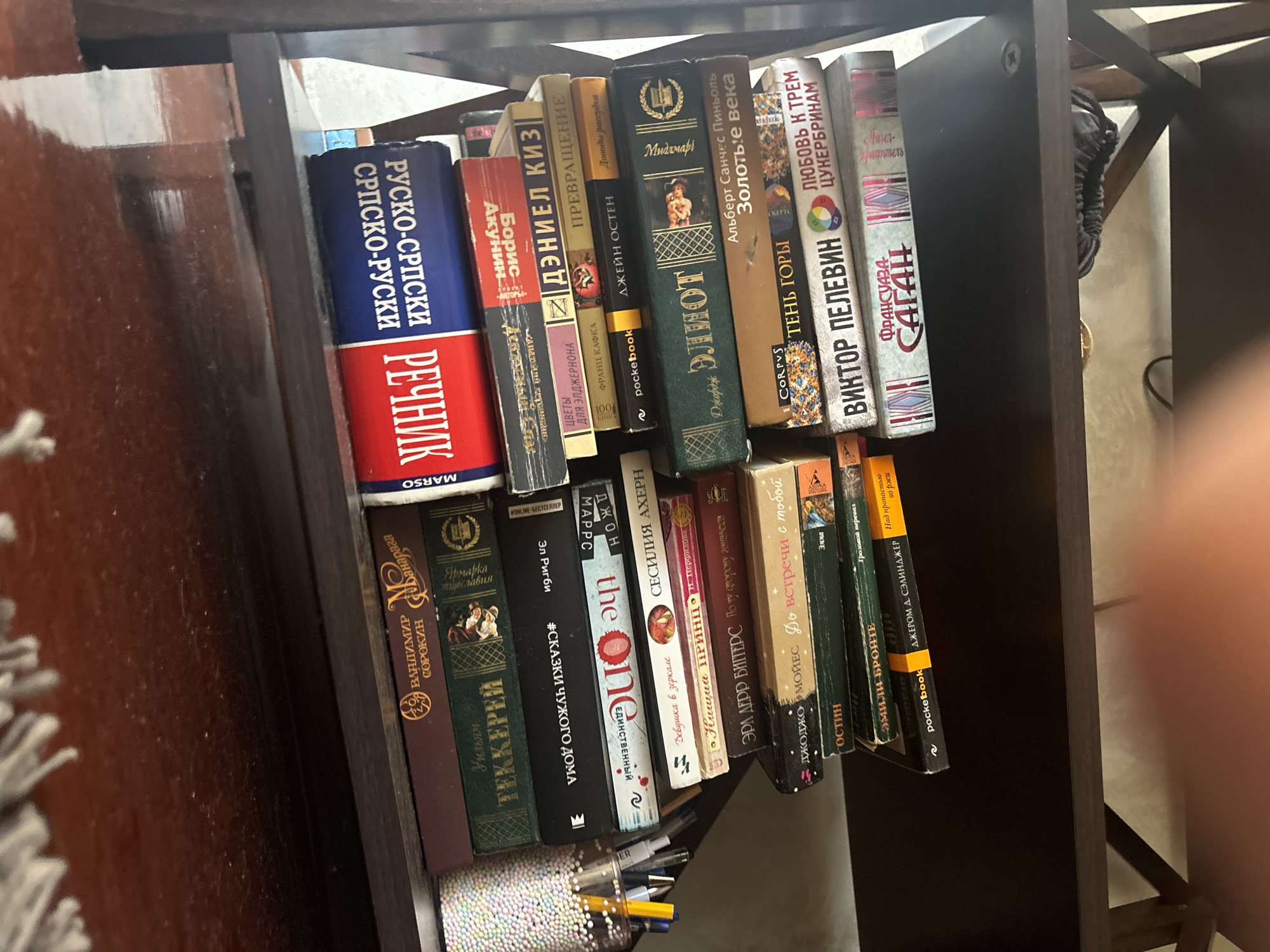 This is a close-up, eye-level shot of a dark wood bookshelf laden with books. The books are stacked in multiple rows, some lying flat and others standing upright. The spines of the books display titles in Cyrillic and Latin alphabets, suggesting a diverse collection. The shelf appears to be part of a larger furniture piece, possibly a desk or a bookcase, with a dark, polished wood finish. The background is blurred, hinting at an indoor environment. The books are the primary focus, with their varied colors and sizes creating a textured visual. The lighting suggests an indoor setting, likely during daylight hours. There are no people or animals visible in the image. The scene evokes a sense of a personal library or a study space. There is no information available to pinpoint the location as Budva, Montenegro.