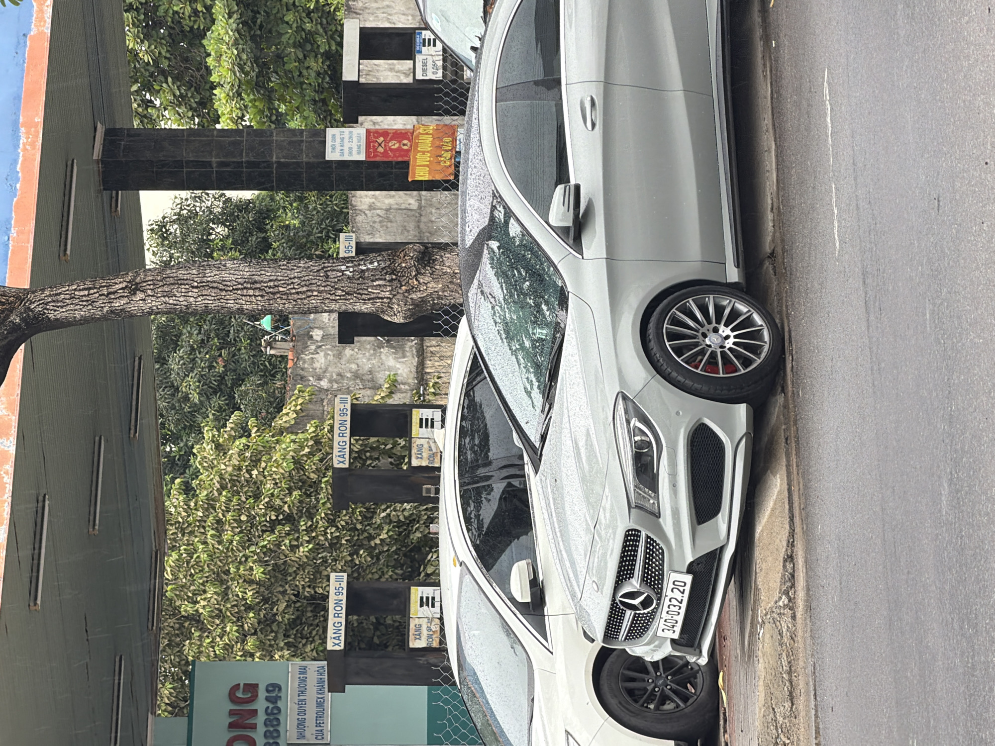 This is a medium shot, eye-level photograph taken in Nha Trang, Vietnam. The scene depicts a rainy day at a gas station with two cars parked. In the foreground, a silver Mercedes-Benz CLA coupe is prominently displayed, its body speckled with raindrops. The car has a license plate that reads "34D-032.20". Next to it, a white sedan, possibly a Ford Focus, is partially visible on the left. The gas station features multiple fuel pumps, with signs indicating "XĂNG RON 95-III" (RON 95-III gasoline) and "DIESEL". A large tree with rough bark stands to the left of the Mercedes, with its canopy extending over the gas station canopy. Behind the cars, a building with black tiled pillars and signage is visible. One sign includes Vietnamese text and a red "X" symbol, possibly indicating a restriction. Another sign in orange reads "KHU VUC QUAN SU CAM VAO" (Military Area, No Entry). The ground is wet, reflecting the overcast sky and the rain. The overall atmosphere is that of a typical urban scene in Vietnam during a downpour.
