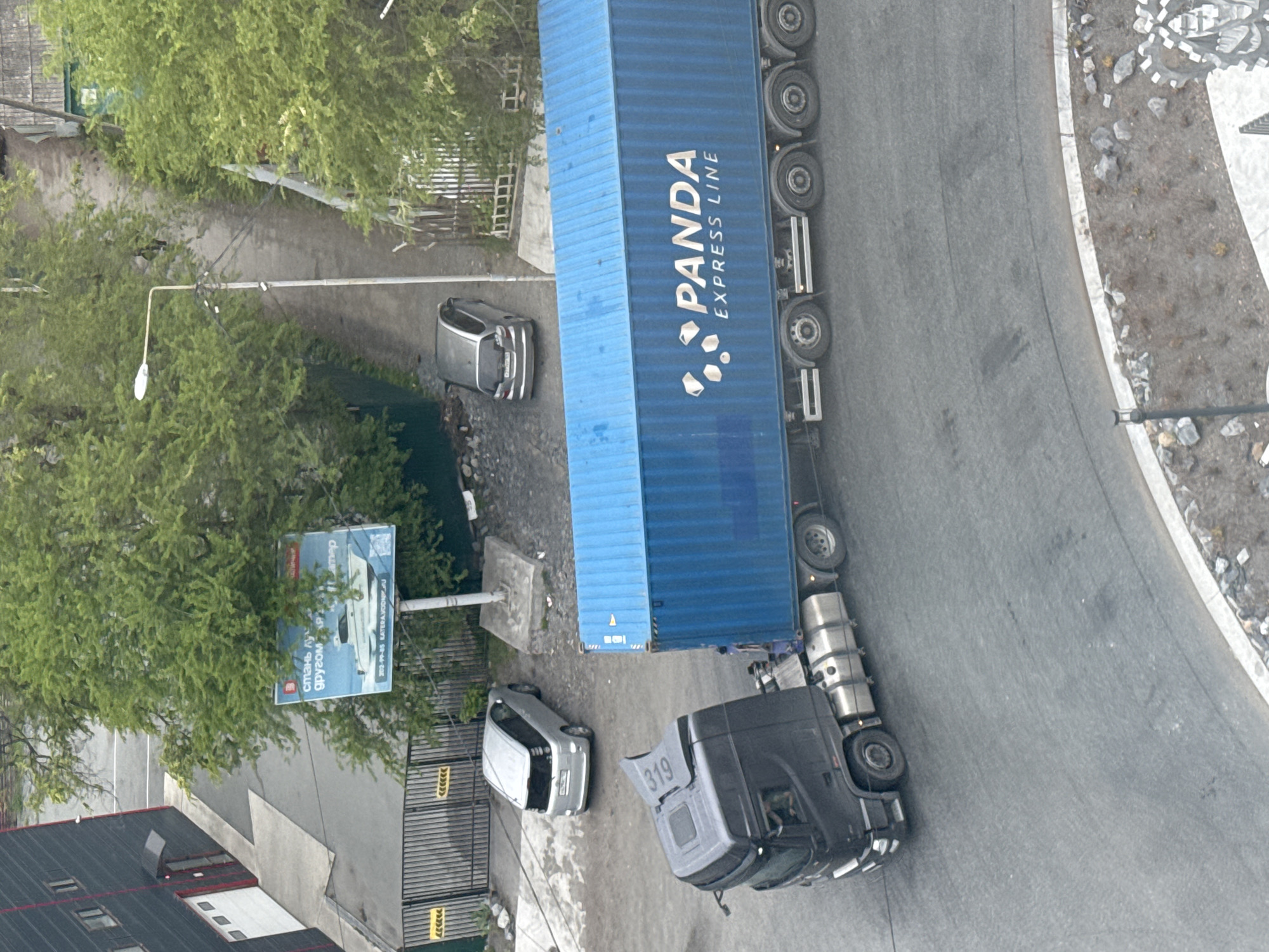 The image, taken from an elevated perspective, captures a scene in Vladivostok, Russia. A large, blue shipping container truck bearing the "PANDA EXPRESS LINE" logo occupies a significant portion of the frame, positioned on a grey paved road that curves slightly. The truck's tractor unit is black and displays the number "319" on its roof. Two light grey passenger cars are parked nearby. A green tree, a street lamp, and a billboard are visible on the left of the image. The background includes buildings with roofs of varying colors. The surroundings are mostly urban with some greenery visible. A decorative stone feature is also seen in the background.