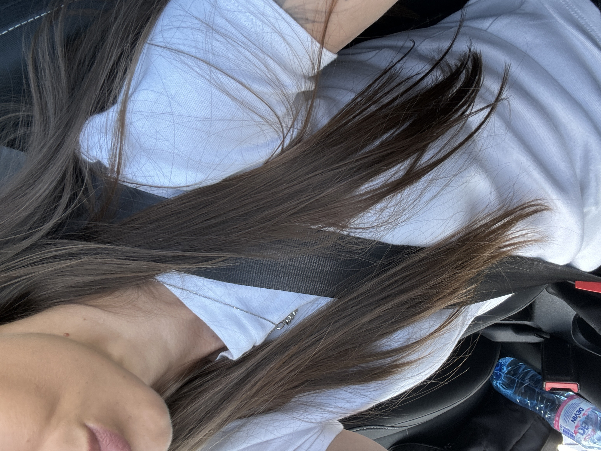 The image shows a close-up of a person wearing a white t-shirt and a seatbelt inside a vehicle. Their long, dark brown hair is visible, framing their face and shoulders. A silver necklace with a pendant is worn around their neck. The background suggests the interior of a car, with black upholstery and a blue water bottle on the floor. The lighting indicates it is daytime, and the image appears to have been taken from a high angle. The setting is not specified as being in Aheloy, Bulgaria.