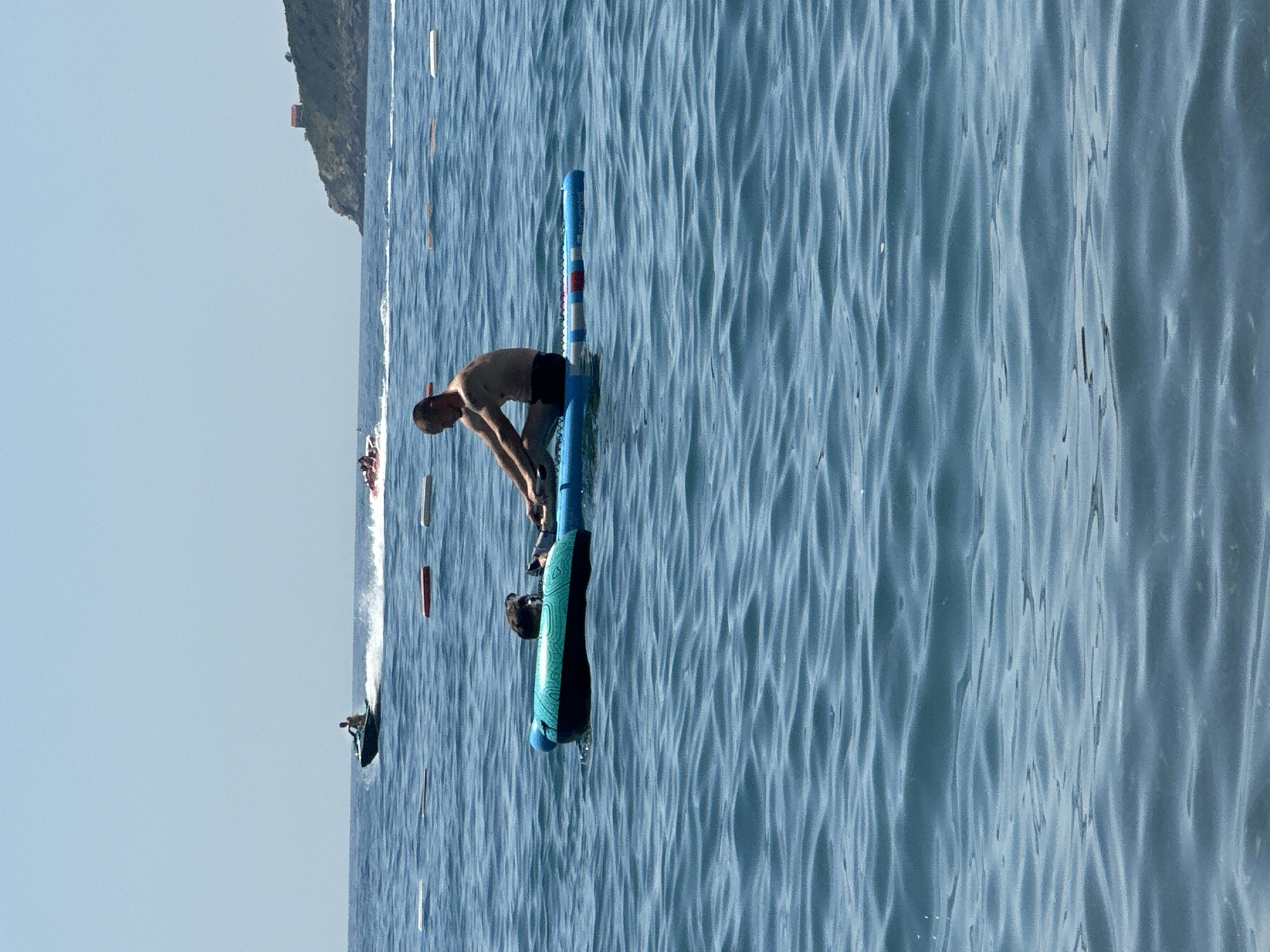 In the city of Boreti, Montenegro, on a clear, sunny day, a man is seated on a blue and red inflatable paddleboard in the calm, blue sea. He is facing forward, with his arms extended as if paddling. Next to him, on the inflatable board, is a dark-colored dog, also facing forward. In the background, the sea stretches out to the horizon under a pale blue sky. Several white and red buoys are visible floating on the water in a line. Further in the distance, a jet ski with a rider is creating a white wake as it moves across the water. Another, smaller jet ski is further out. On the right side of the image, a green, hilly coastline with a small, red-roofed building is visible. The overall scene depicts a leisurely day at the beach with recreational activities taking place on the water.