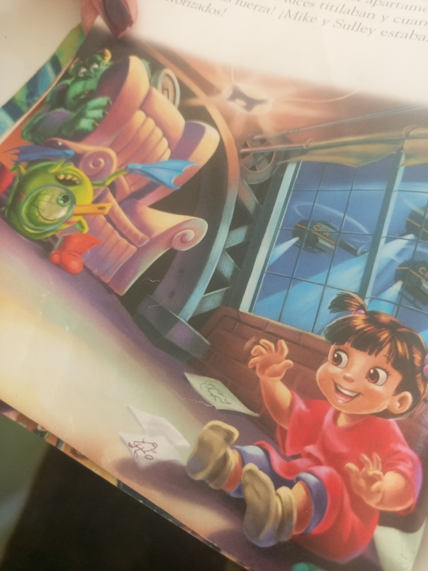 The image is a photograph of an open book, featuring an illustration from the Disney movie "Monsters, Inc." In the foreground, a young girl, identified as Boo, is depicted sitting and smiling with her hands raised, wearing a red dress and light-colored boots. Behind her, there are elements of a factory setting, including large pipes and windows that show stylized vehicles flying outside. To the left of Boo, two cartoon monsters are visible: one is green and purple with a large eye and wings, and another is a larger, green, furry creature in the background. The overall scene is colorful and whimsical, characteristic of children's book illustrations. There is no discernible location within the city of La Tagua, Venezuela, or any other real-world geographical setting depicted in this image; it is purely illustrative and fantastical.