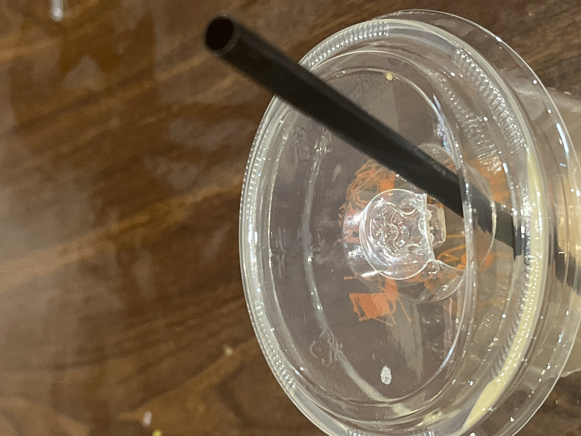 A close-up, overhead shot captures a black plastic straw inserted into a clear plastic cup, likely containing a beverage with a yellowish hue and some foamy residue. The cup is sealed with a clear plastic lid, which has a small, molded plastic tab for opening. The lid exhibits some condensation and smudges. The background is a dark, polished wooden surface, suggesting it might be a table. No people or animals are visible in the image. The scene is static and appears to be indoors. The specific location in Tanjung Balai Karimun, Indonesia, is not discernible from the visual cues provided.