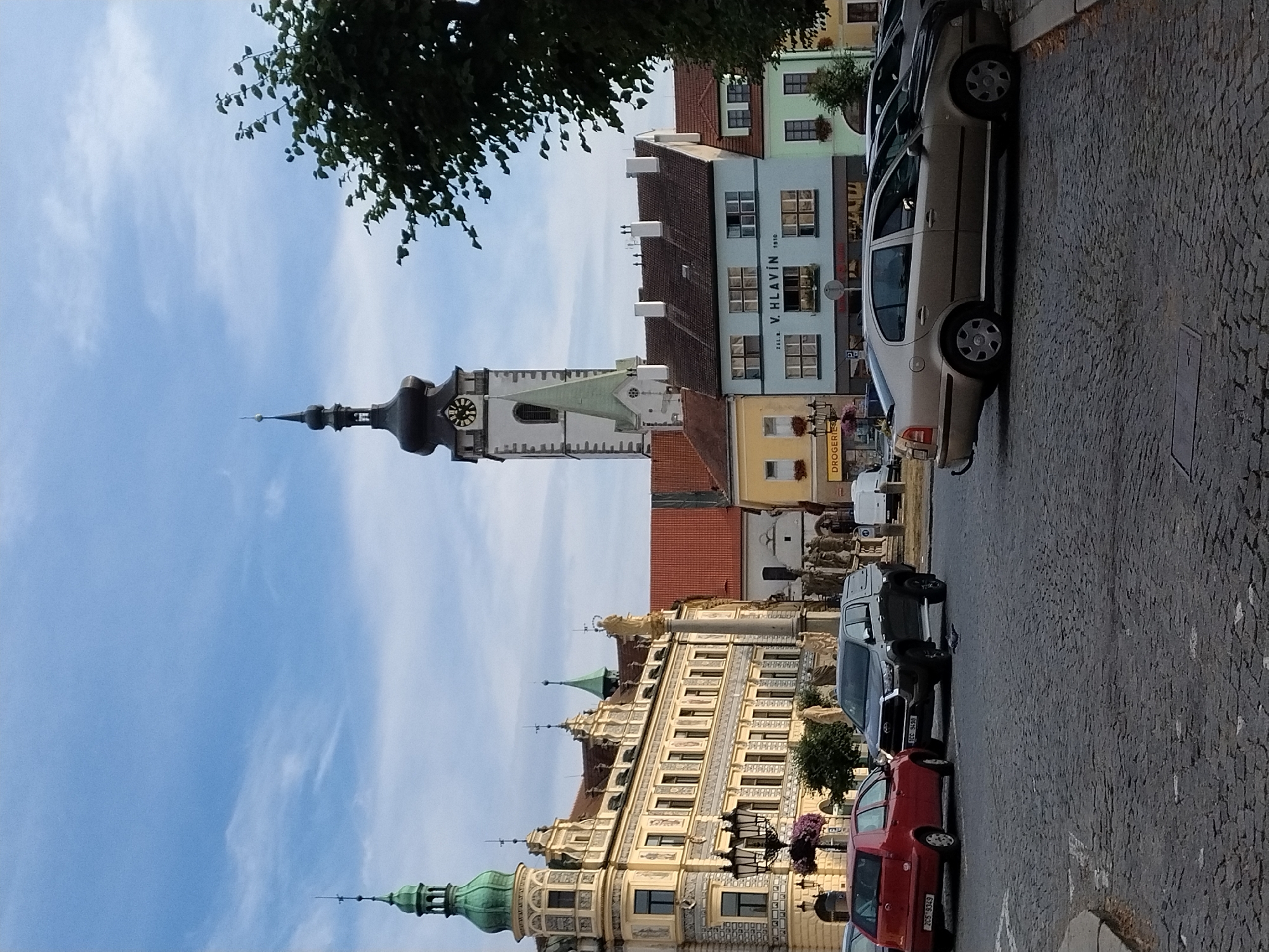 The image shows a view of the Great Square (Velké náměstí) in Písek, Czechia. The square is paved with cobblestones and features a variety of buildings and objects. Dominating the background is the Church of the Nativity of the Virgin Mary (Kostel Narození Panny Marie) with its distinctive tower and spire. To the left of the church is a large, ornate yellow building with green spires and numerous windows. In front of this building is a street lamp with hanging flower baskets. In the middle ground, several cars are parked on the square, including a red hatchback, a dark-colored pickup truck with a canopy, and a light-colored sedan. Other buildings, including a pale blue structure with the text "V. HLAVÍN 1910" and a green building, are visible. A statue on a tall pedestal stands near the center of the square. The sky is partly cloudy.