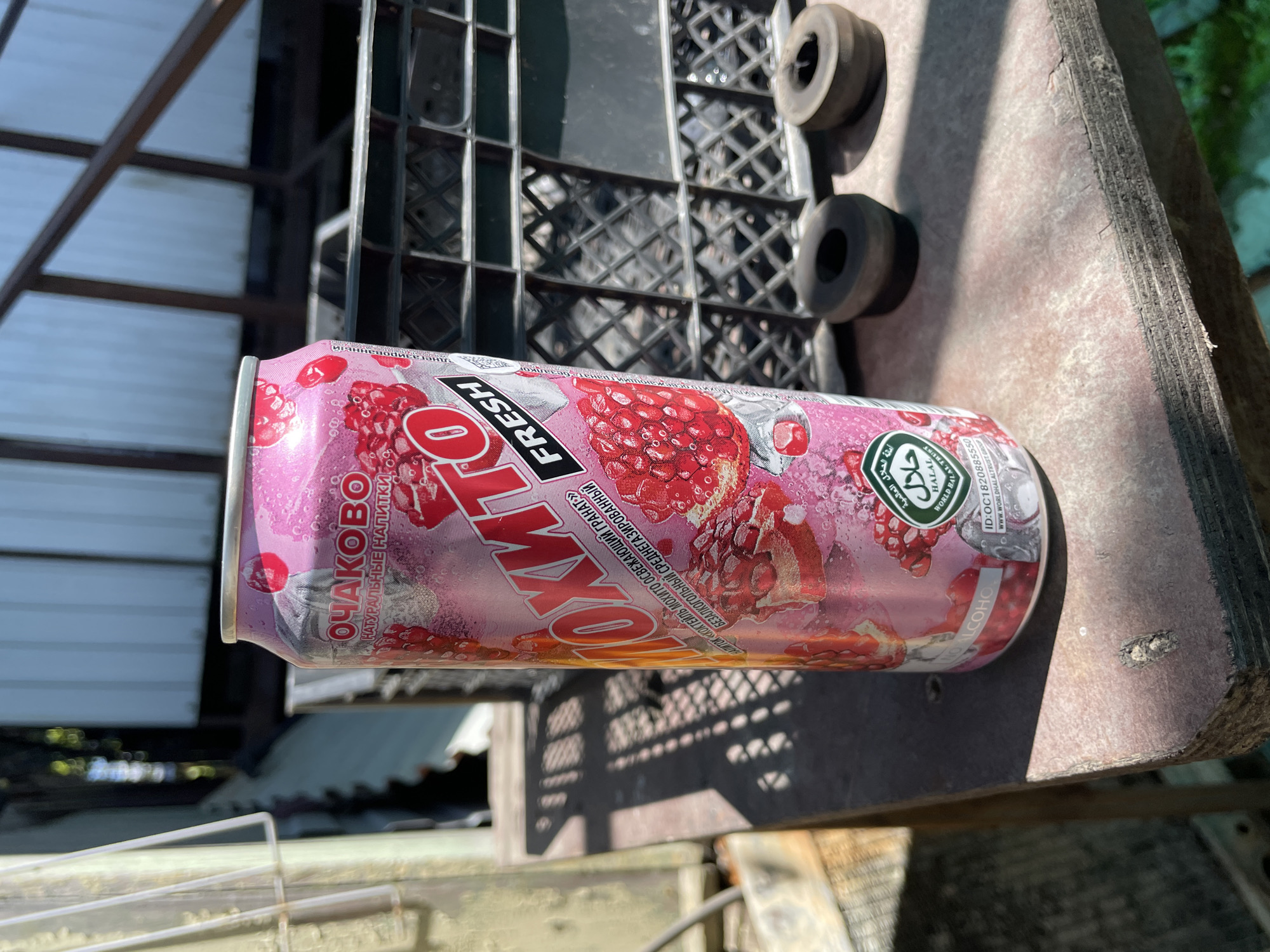 A can of "Ohako Mohito" refreshing pomegranate cocktail drink, manufactured by the Ohako company (Очаково) - a Russian beverage company, is seen on top of a weathered, horizontal, wood surface. The can is pink, printed with images of pomegranates. The Russian word "ОЧАКОВО" is vertically printed near the top. The word "FRESH" appears above the word "MOHITO." The can also features the Halal symbol. The surrounding environment includes a black plastic grid structure and two black rubber wheels. The scene is partly shaded, with glimpses of a partially-covered outdoor area. There is no clear indication that this scene is located in Olginka, Russia.