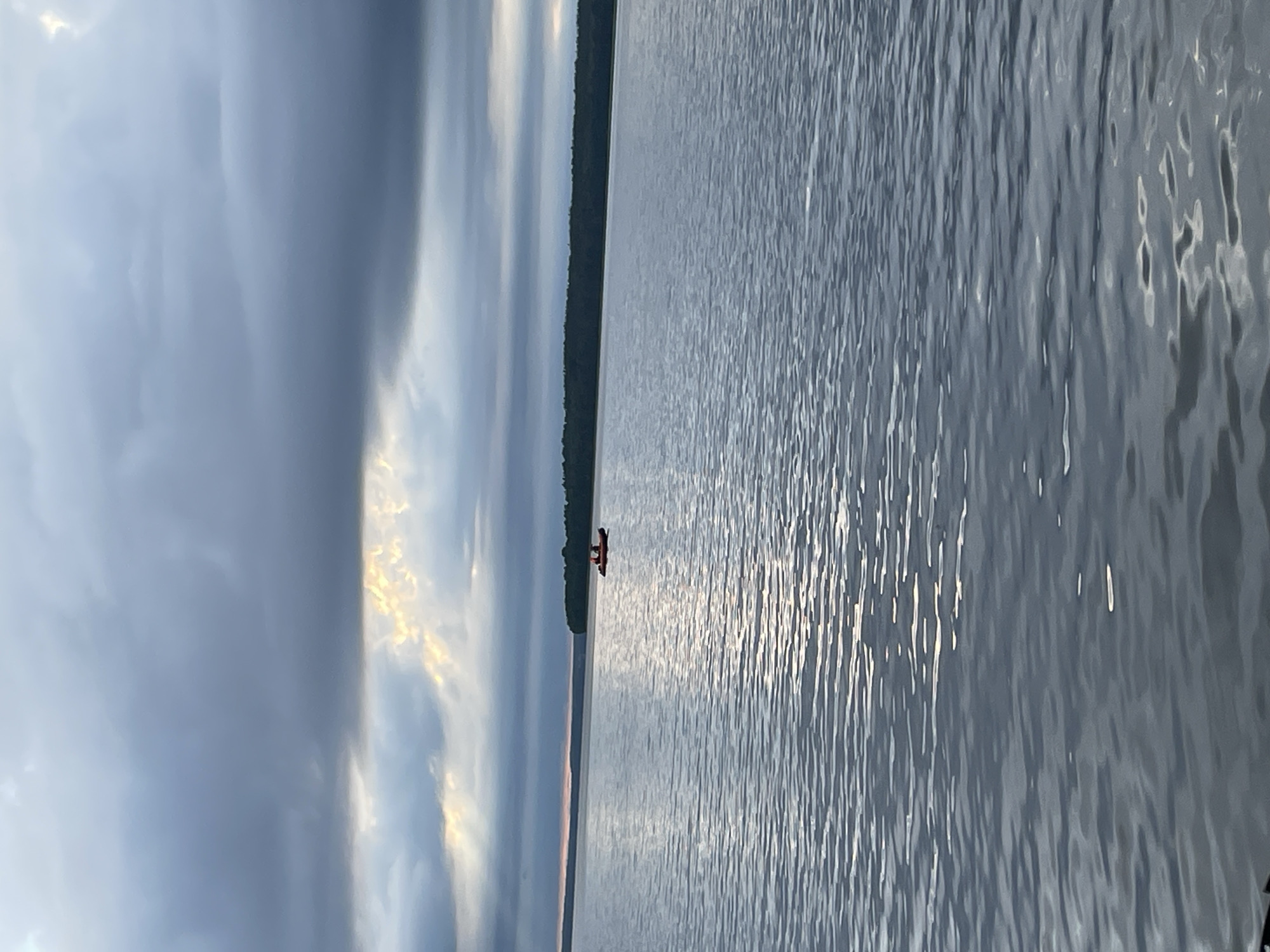 An image captures a serene body of water under a cloudy sky, with a distant treeline on the horizon. In the middle of the water, a small orange boat with two people is visible. The water surface reflects the cloudy sky and sunlight breaking through the clouds. The scene appears to be a calm day on a lake or river, possibly in Yablonovka, Russia, given the natural landscape.