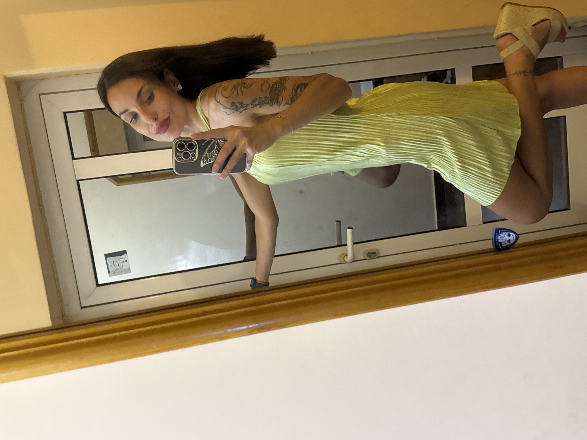 A full-body, mirror selfie shows a woman posing with her right leg bent and extended behind her. She is wearing a lime green, pleated slip dress, wedge sandals, and has dark hair. Tattoos are visible on her left arm. The background appears to be a doorway with a mirror on the left, reflecting the woman and a portion of a room with an orange-toned wall. There are stickers on the door frame, one of which resembles Groot from Guardians of the Galaxy. The image was taken indoors, likely in a residential setting. The location is not specified in the image or provided context.