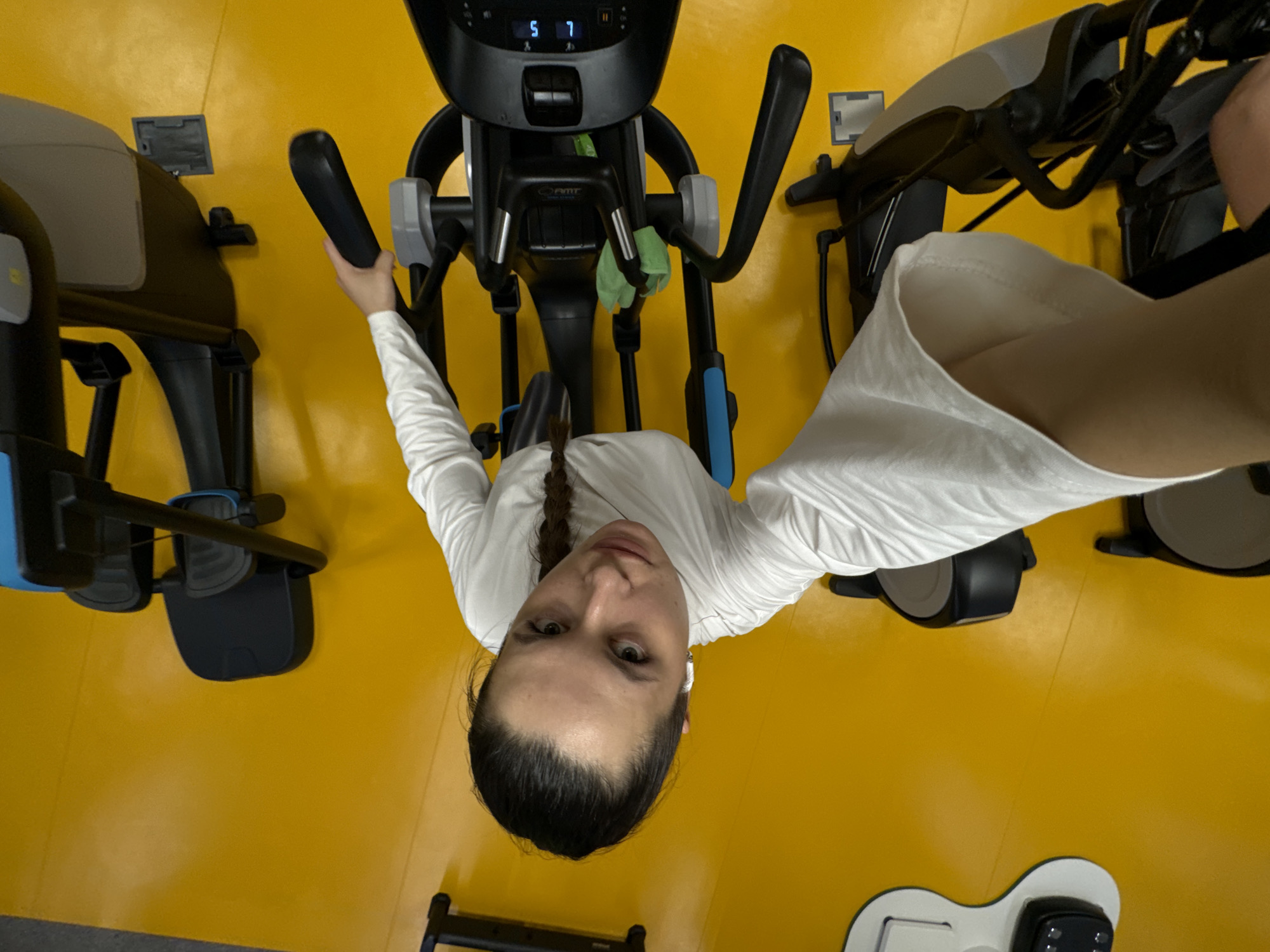 This top-down image captures a woman exercising in what appears to be an indoor gym or fitness center in Lesnoi Gorodok, Russia. The striking feature of the setting is the bright, uniform yellow color of the floor and partially visible walls.
The main subject is a woman with dark hair styled in a braid, wearing a white long-sleeved top. She is positioned on an exercise machine, likely an elliptical trainer, and looks directly at the camera with a neutral expression. She has small white wireless earbuds in her ears.
The scene features multiple exercise machines. The woman is on a black and gray elliptical trainer with blue accents, holding its handlebars. A small green towel is draped over the machine's front handlebars. The digital display on the machine's console shows the numbers "5 7". Below the console, the text "PMT" is visible on the machine's frame. Portions of two other similar elliptical machines are visible on the left and right sides of the frame, indicating a row of equipment. In the bottom right corner, a white and black object with controls, possibly part of another machine or fitness accessory, is partially visible on the yellow floor.
The activity depicted is a workout session, with the woman either actively exercising or paused on the elliptical machine. The overall impression is that of a contemporary and functional fitness environment.