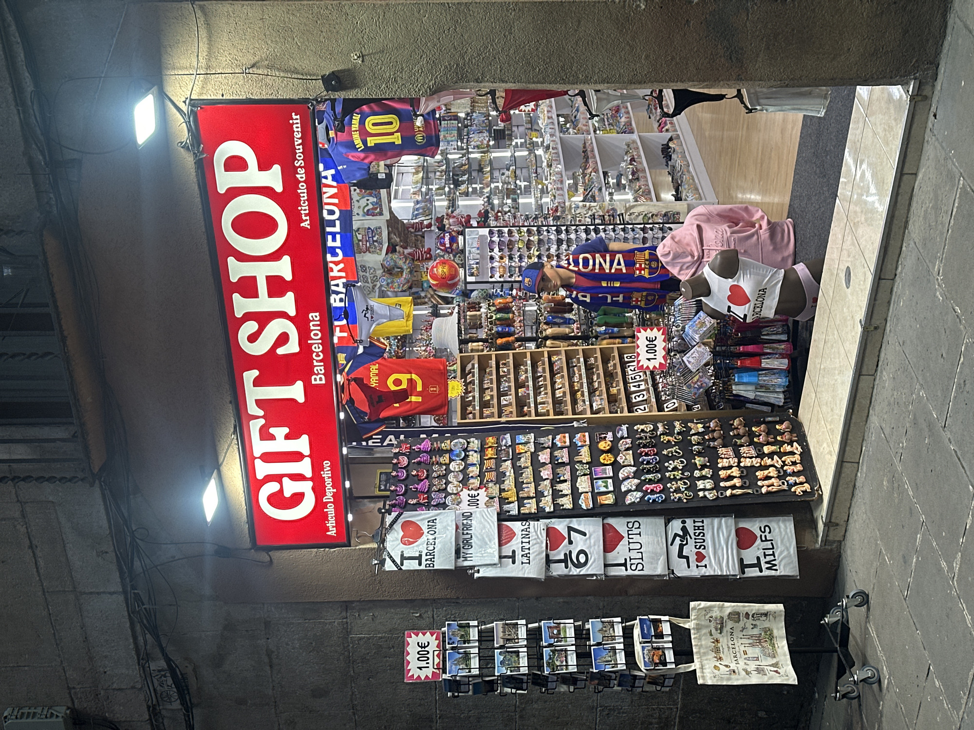The image depicts a nighttime street view of a brightly lit souvenir shop in Barcelona, Spain, identified by a prominent red sign reading "GIFT SHOP Barcelona" and "Articulo de Souvenir." The shop displays a variety of merchandise, including t-shirts with slogans like "I LOVE BARCELONA," "I LOVE LATINAS," "I LOVE SLUTS," "I LOVE SUSHI," and "I LOVE MILFS." Football jerseys, including one with the name "LAMINE YAMAL 10," are also visible. Magnets and small souvenirs are arranged on racks near the entrance. A mannequin wearing an "I LOVE BARCELONA" crop top and underwear is positioned in the shop window, accompanied by another mannequin wearing a pink hooded sweatshirt. Outside the shop, a rack of postcards and a tote bag with Barcelona landmarks are displayed. The street and sidewalk appear to be made of stone. The overall scene suggests a commercial area focused on tourism and novelty items.
