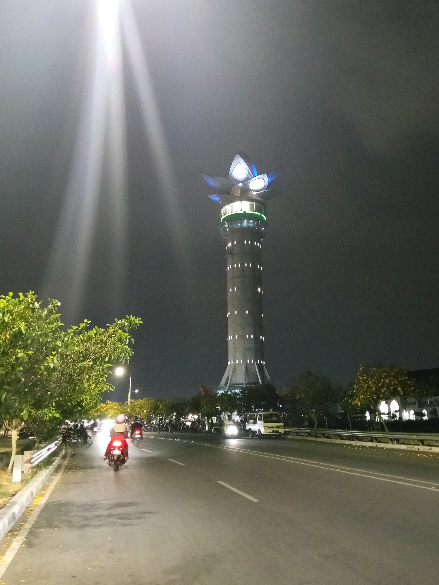 In Purwokerto, Indonesia, at night, a tall, cylindrical tower with a lotus-shaped, illuminated top stands prominently against a dark sky. The tower is dotted with small, white lights. In the foreground, a road stretches out, with a few motorcycles and a small truck visible. On the left side of the road, there are trees and a sidewalk, with a streetlamp providing illumination. The sky is mostly dark, but a bright beam of light shines down from the upper left, illuminating a portion of the sky and the tops of the trees. The overall scene suggests a nighttime urban environment with a notable landmark.