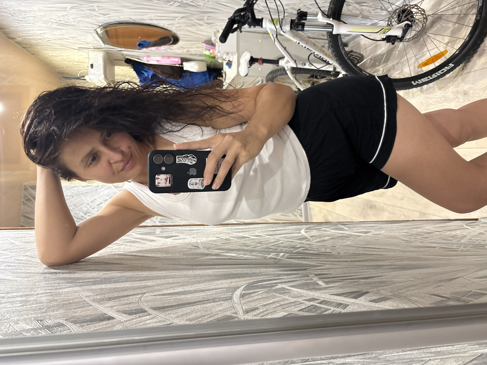 A woman with long, dark, wet hair is taking a selfie in a hallway. She is wearing a white tank top and black shorts. She is leaning against a textured, silver and white wall, with her right arm bent and her hand resting on her head. Her left arm is holding a black smartphone, which is angled to capture her reflection. A bicycle with a white frame and black tires is visible behind her, leaning against the wall. The hallway appears to be part of an apartment building, with light-colored patterned wallpaper and a wooden floor. A round mirror is hanging on the wall in the background, reflecting a blue object and other items on a shelf. The overall environment suggests an indoor residential setting.