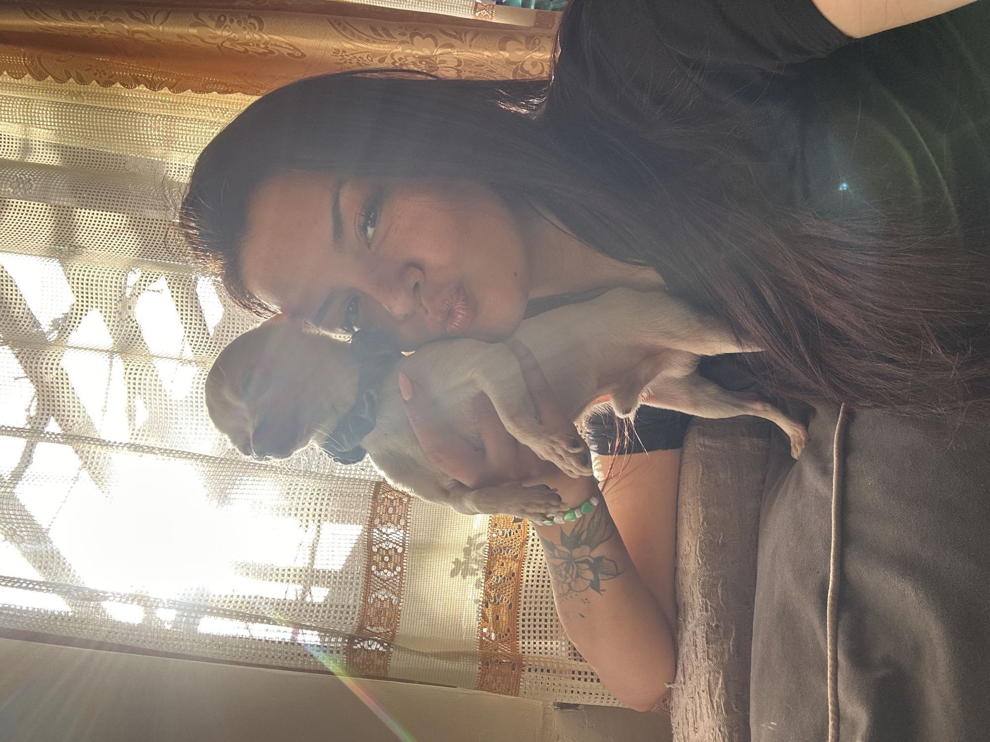 A woman with long, dark hair and a flower tattoo on her left arm is holding a small, light-colored dog. They are positioned indoors next to a window covered with a lace curtain, possibly in a domestic setting. The location is not discernible in the image.